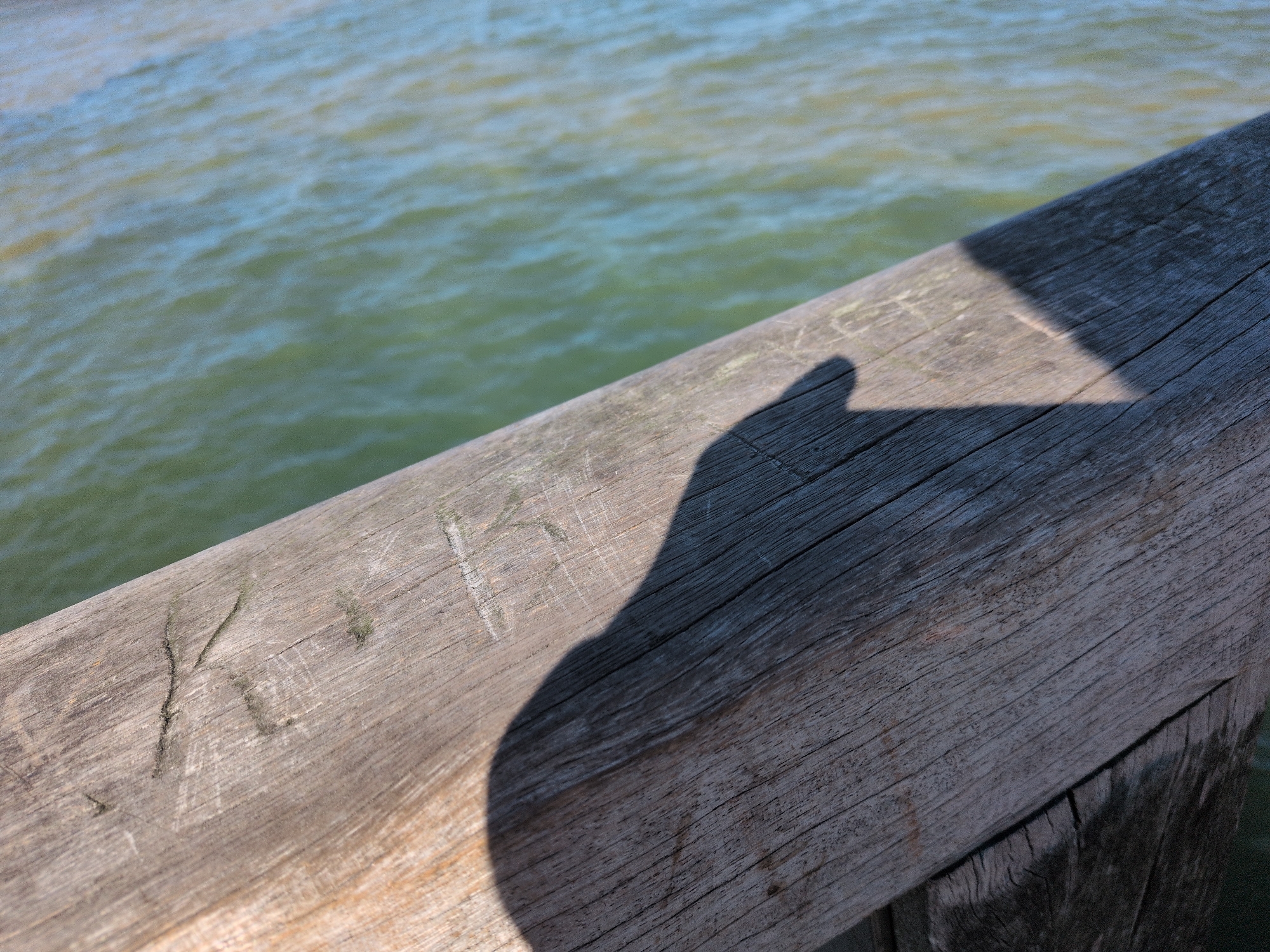 The image is a close-up, eye-level shot of a weathered wooden railing, casting a shadow of a person's head and upper body. The railing is positioned diagonally across the frame, with the shadow falling from the upper right to the lower left. The wood grain is visible, and there are faint markings or carvings on its surface. Behind the railing, the sunlit surface of the Baltic Sea is visible, appearing choppy and greenish-blue. The scene suggests a person is standing on a pier or boardwalk in Kühlungsborn, Germany, looking out at the sea, with their shadow cast upon the railing.