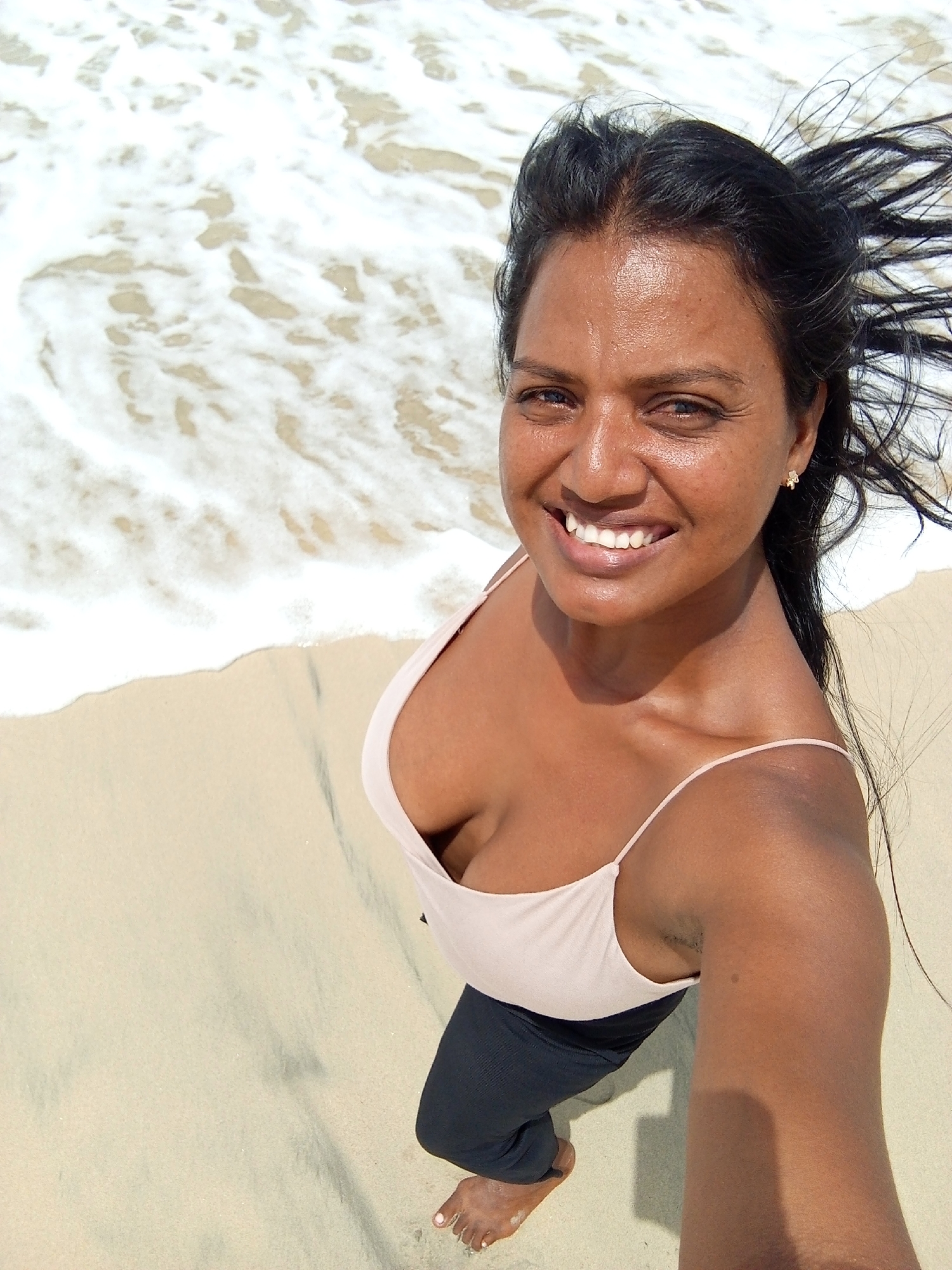 A woman is taking a selfie at a beach in Altagracia, Venezuela. She is fair-skinned with dark hair, wearing a light pink tank top and dark pants. The ocean is visible in the background with white foamy waves lapping on the sand. The sand is light tan. The woman is smiling and appears to be enjoying herself.