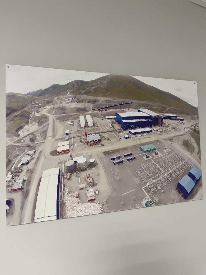 This image depicts an aerial view of a large-scale industrial complex located in the city of Im. Gastello, Russia. The complex is situated within a remote, hilly landscape characterized by barren, rocky terrain and sparse green vegetation under an overcast sky.
The scene reveals a network of industrial infrastructure, including multiple large blue buildings centrally located, indicative of primary processing or plant facilities. A prominent conveyor belt system extends from these central buildings, ascending into the background hills, suggesting material transport from an excavation or mining area.
To the right, a substantial electrical substation is visible, equipped with numerous gantries, transformers, and power lines, indicating significant energy distribution. Near the foreground-right, several small blue arched storage structures are present.
In the mid-ground, a cluster of silver cylindrical storage tanks, likely for liquids, is situated alongside various smaller support buildings and equipment. A distinct red and yellow building with an adjacent red lattice-boom crane suggests active construction or maintenance operations. Towards the foreground-left, a large white arched structure, possibly a warehouse, stands next to a significant accumulation of white bags or sacks, indicating bulk material storage. Numerous other smaller buildings, temporary structures, and equipment are distributed throughout the site.
The ground exhibits extensive signs of disturbance, grading, and excavation, consistent with ongoing construction and industrial activity. A network of dirt and gravel roads connects the various sections of the complex. No human figures are discernible in the image.