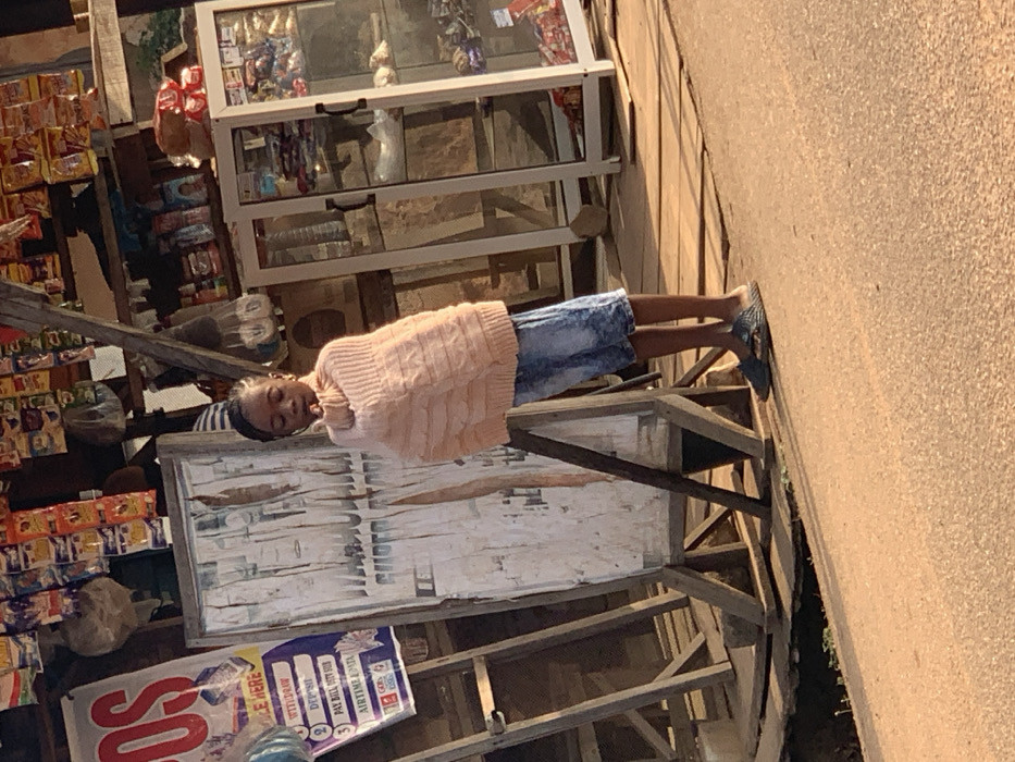 A child, wearing a pink sweater and denim shorts, lies asleep on what appears to be a wooden structure outside a roadside shop. The shop displays various goods, including snacks and drinks. Adjacent to the wooden structure is a sign with text in English including, "PLACE HERE," "WITHDRAW," "DEPOSIT," "PAY BILL," and "AIRTIME." The image is tilted, showcasing the street and the exterior of the commercial space.