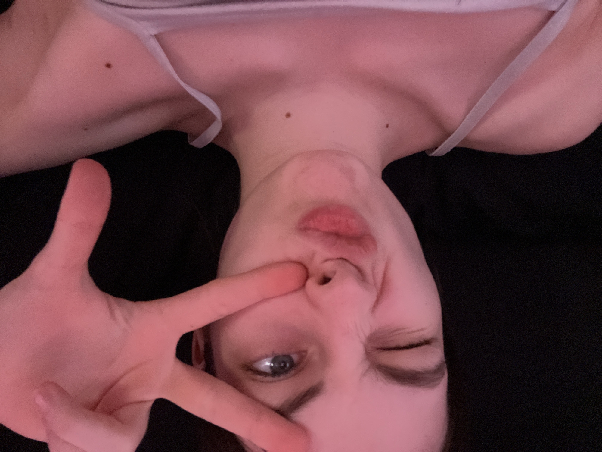 A woman is lying down on a black surface and taking a selfie. She has fair skin, one blue eye visible, and is winking. Her mouth is pursed, and she is making a peace sign with her hand in front of her face. She is wearing a light-colored top with thin straps. The lighting has a pinkish hue. There are two moles visible on her neck. The location is not identifiable.