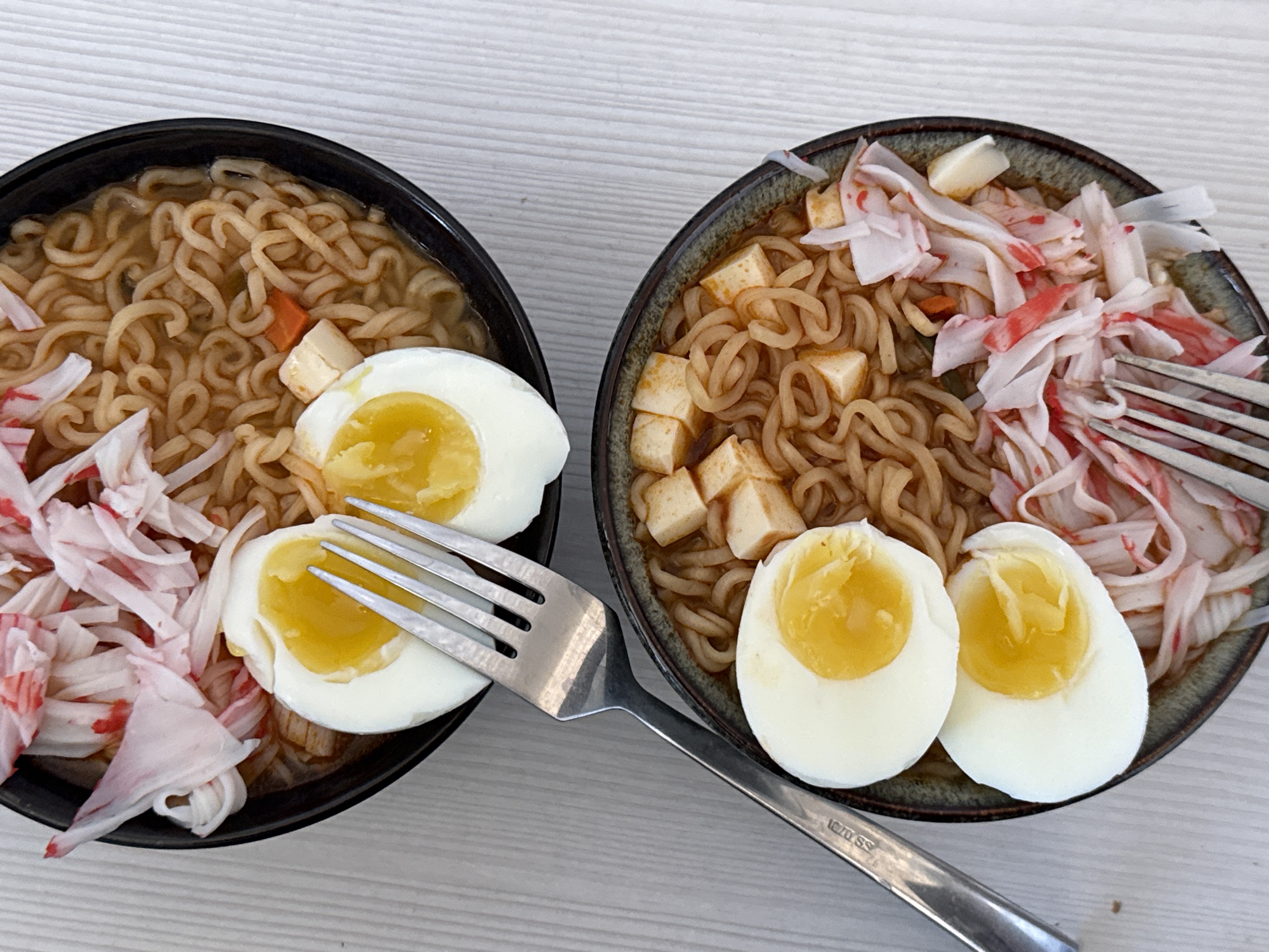 Two bowls of ramen are on a light gray surface. The left bowl is black and contains noodles, imitation crab meat, tofu and two halves of a soft-boiled egg. The right bowl is dark and contains noodles, imitation crab meat, tofu and two halves of a soft-boiled egg. A fork lies between the bowls. The scene is well lit. The location is not discernible from the image.