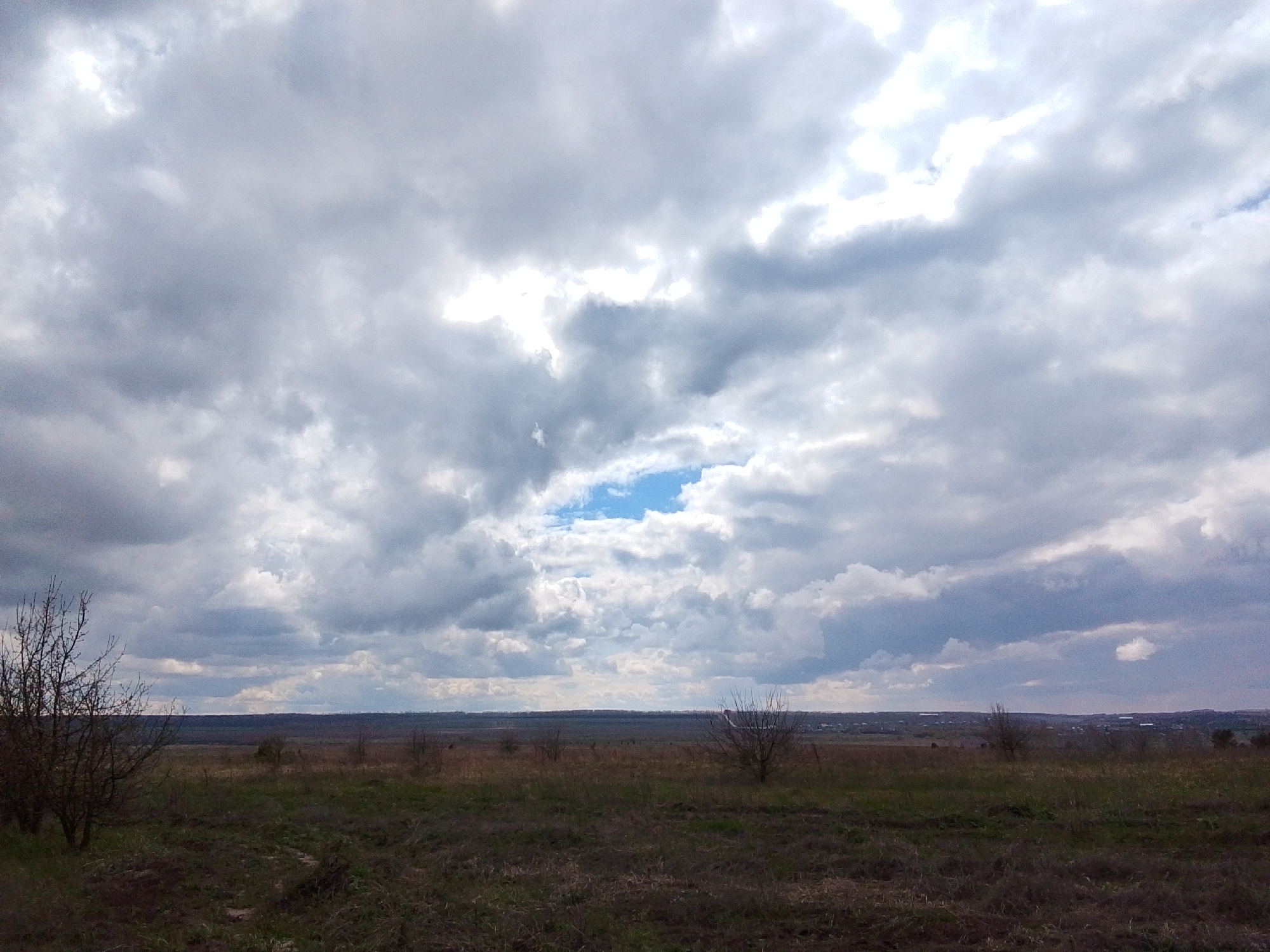 The image presents a wide, expansive landscape under a dramatic, cloudy sky, likely depicting a rural area near Khlevnoye, Russia. The scene is dominated by a vast sky filled with heavy grey and white clouds, suggesting an overcast day. A distinct patch of bright blue sky is visible towards the center, hinting at the clouds breaking or a brief moment of sunlight.
The foreground and midground consist of a large, open field or undeveloped land, covered predominantly with dry, brown, and green-brown grass and patchy, sparse vegetation. Several leafless trees or bushes are scattered across the landscape, particularly a cluster on the left side and a few smaller ones in the midground and right. Their bare branches indicate either late winter or early spring.
In the far distance, a subtle horizon line reveals what appears to be a low-lying forest, hills, or a distant settlement with some faint structures. No people or specific objects are visible, emphasizing the natural, untamed aspect of the setting. The weather appears cool and potentially damp due to the cloud cover. The overall impression is one of quiet solitude and vastness under a changing sky. No text is visible in the image.