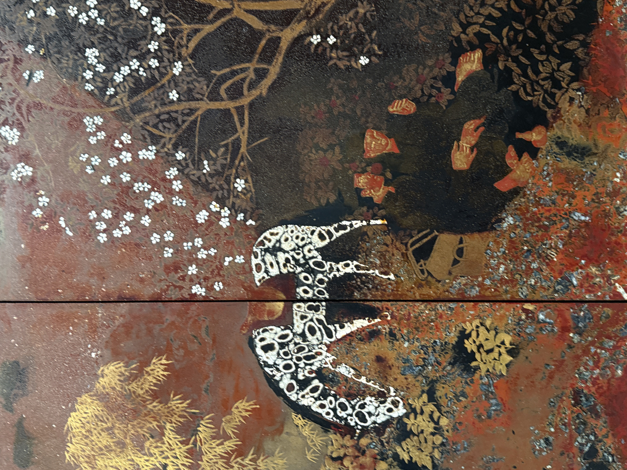 The image depicts a detailed close-up of a decorative screen, likely a Japanese lacquerware screen (byobu). The screen is divided vertically into two panels. The left panel features a large, stylized animal, possibly a horse or deer, with a striking pattern of black and white irregular spots. To the left of this animal is a clump of golden bamboo leaves rendered with fine detail and a metallic sheen. The background of this panel is a mottled reddish-brown.
The right panel displays a dark, textured background against which various elements are depicted. At the top and extending into the middle ground are branches of a tree adorned with small, white, five-petaled flowers, scattered sparsely. Some of these flowers are also present in the upper left corner of the right panel, overlapping the vertical divide. In the lower right section of this panel, a dense cluster of dark foliage is visible, with hints of red and orange tones at the very bottom edge, suggesting fallen leaves or flowers. In the center of the right panel, scattered amongst the dark foliage, are several small, stylized figures rendered in gold. These figures appear to be abstract representations of hands, faces, and possibly objects like a small vase or bottle.
The overall aesthetic is rich and ornate, with a strong emphasis on pattern and texture. The colors are predominantly earthy tones of red, brown, and gold, contrasted with the stark black and white of the spotted animal and the white of the blossoms. The surface of the screen appears to have a granular texture, likely due to the application of lacquer and possibly crushed eggshell or other materials for decorative effect. The lighting suggests a controlled, possibly indoor environment, highlighting the intricate details and reflective qualities of the artwork.
This image was captured in the city of Thủ Đức, Vietnam, though the specific location within the city is not identifiable from the image itself. The artwork is likely a decorative object within a home, gallery, or museum.