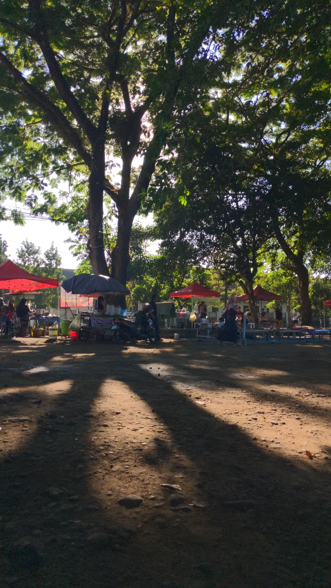 This is a ground-level, outdoor photograph of a market or food stall area situated in Magelang, Indonesia. The scene is dominated by large trees casting dappled shadows across a sandy, uneven ground. In the background, several stalls are set up under red and dark umbrellas. Some of these stalls appear to be selling food, with tables and various items displayed. People are present, some standing and interacting at the stalls, while others are seated. Motorcycles are parked near some of the stalls. The lighting suggests it is daytime, with sunlight filtering through the tree canopy. The overall atmosphere appears casual and typical of a local market setting.