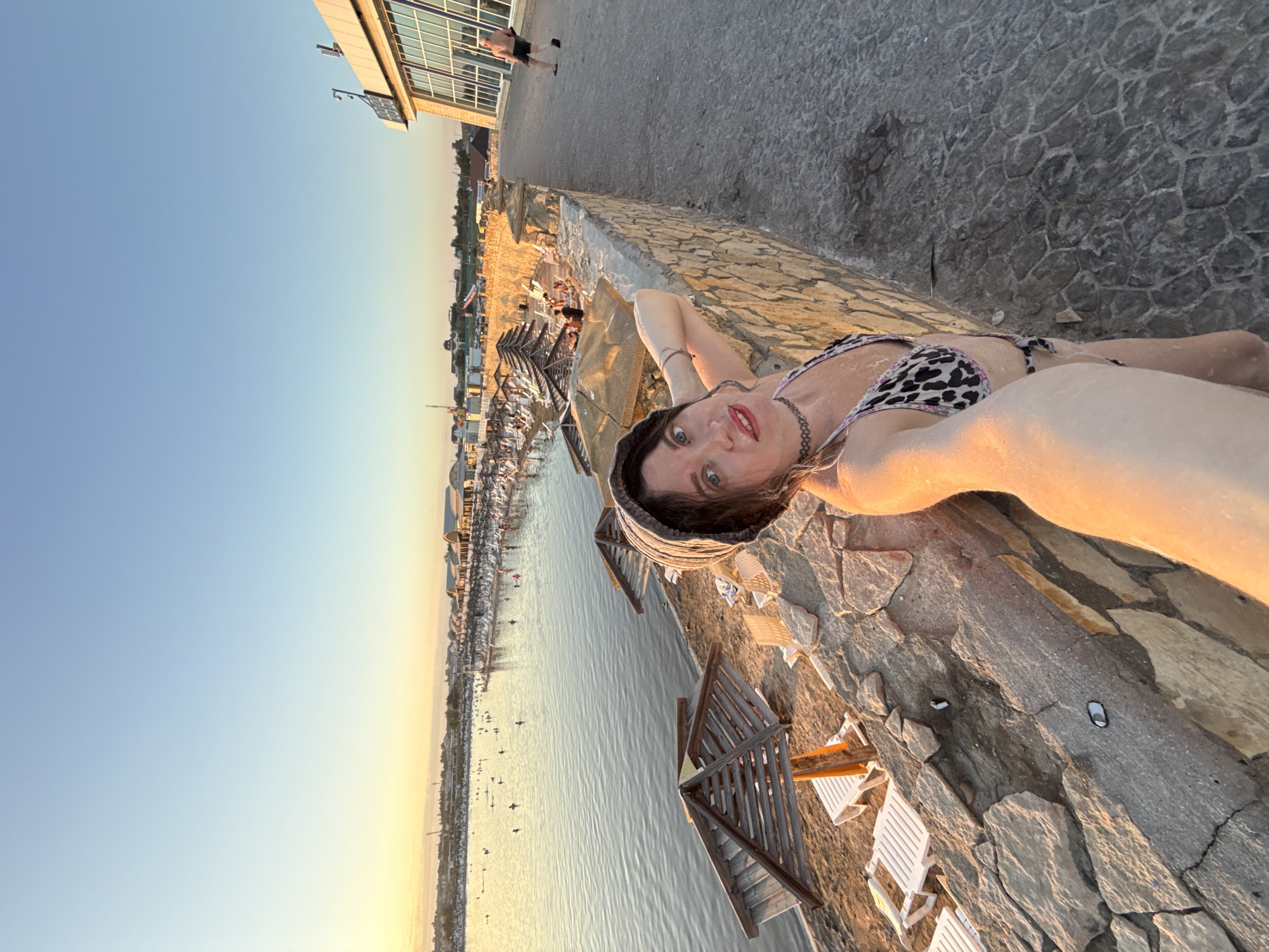 An outdoor, wide-angle, selfie-style photograph taken at sunset on the shore of the salt lake in Sol-Iletsk, Russia. The woman holding the camera is positioned in the foreground, her right arm raised to capture the shot. She is wearing a leopard-print bikini and a dark headscarf. Her gaze is directed at the camera. Behind her, a stone retaining wall runs along the edge of a paved walkway. To the left of the wall, a beach with lounge chairs and wooden umbrellas is visible, bordering a calm body of water populated by numerous people. In the background, a strip of land with buildings and vegetation stretches across the horizon, under a clear, pale blue sky that transitions to yellow at the horizon. To the right, a modern, glass-fronted building is partially visible. A man in black shorts is walking away from the camera on the walkway. The lighting suggests the golden hour, with warm tones illuminating the scene.