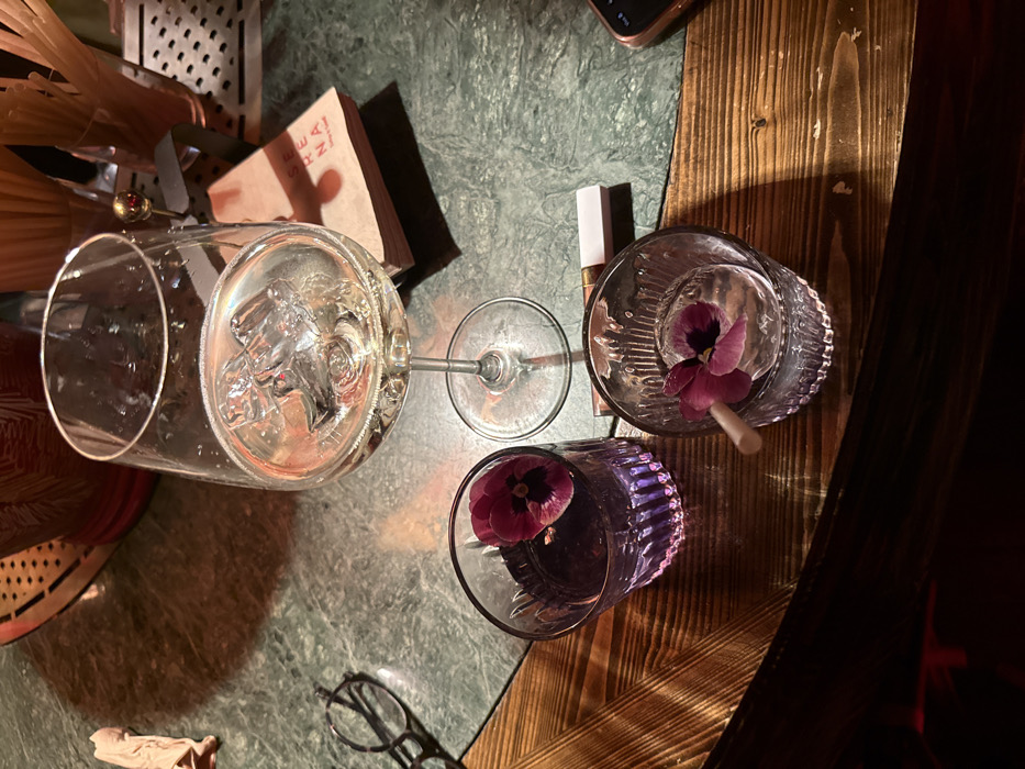 The scene is an interior shot of a table or bar counter at Serena Cafe & Wine, located at 37 Abovyan Street, Yerevan, Armenia. Three prepared drinks are centrally positioned. A clear stemmed wine glass, containing a pale yellow liquid and several ice cubes, rests slightly right of center. To its lower left, a fluted glass holds a purple-colored beverage, garnished with a single purple pansy flower. To the lower right, another fluted glass contains a clear liquid with a large spherical ice cube, also garnished with a purple pansy flower and a light-colored straw.
A pair of dark-framed eyeglasses lies on the green, mottled stone table surface to the left of the purple drink. A white and bronze-toned lipstick or lip gloss tube is situated between the two fluted glasses. A fragment of a rose gold-colored smartphone is visible in the bottom right corner of the frame. The table surfaces consist of the aforementioned green stone on the left and upper areas, and a section of dark, horizontally-grained wood on the lower right, indicative of a bar top. In the background, a stack of small, light-brown paper items branded "SERENA" is visible near the wine glass, alongside containers of slender sticks, likely straws, and a metallic grate, suggesting a bar environment. The lighting is dim and warm, producing shadows and reflections on the glass and liquid surfaces. No individuals are present.