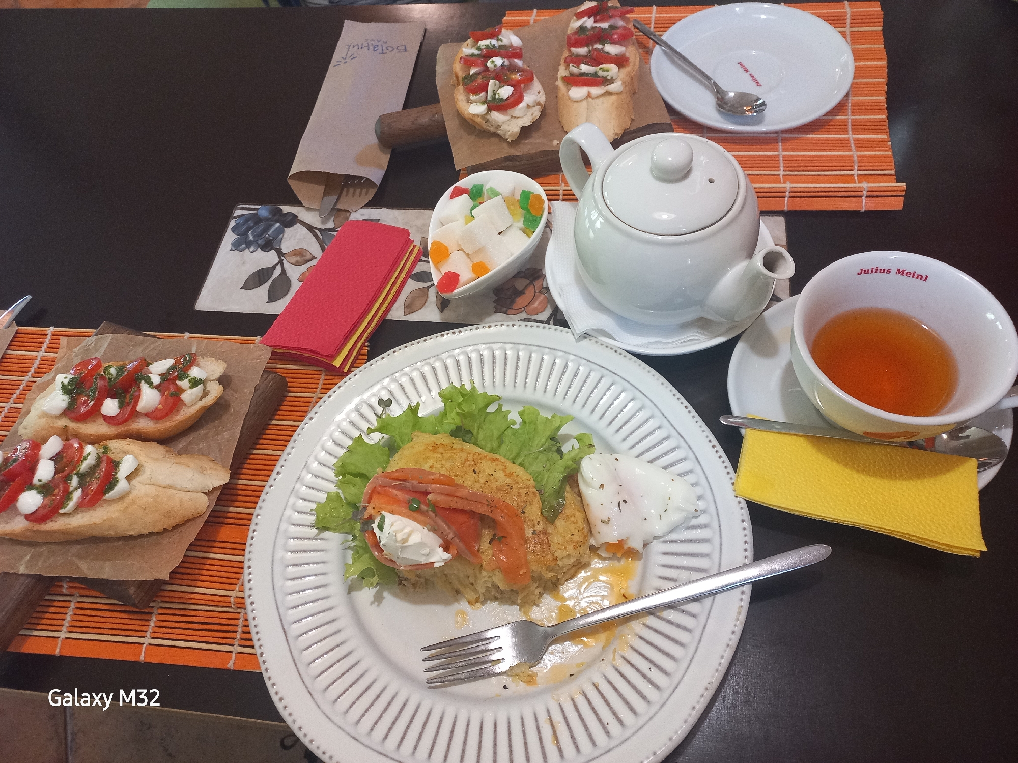 The image shows a table setting in what appears to be a café or restaurant. The setting includes:
* A white plate with a dish consisting of fried bread, lettuce, salmon, soft white cheese, and a poached egg. A fork rests on the plate, and there are traces of food, suggesting the dish has been partially eaten.
* Two pieces of bruschetta on a wooden cutting board covered in parchment paper. They are topped with sliced tomatoes, mozzarella, and possibly basil pesto.
* A white teapot on a saucer.
* A white cup filled with tea, sitting on a saucer. The cup and saucer bear the logo "Julius Meinl".
* A small bowl of candied fruit.
* A stack of red napkins.
* A small orange bamboo placemat with a white plate on it.
The camera model "Galaxy M32" is mentioned in the bottom left corner of the picture. The overall impression is of a leisurely meal in a pleasant setting, with a focus on presentation and variety of food items. Given the products displayed, the location could be considered to be any cafe around the globe that has a Julius Meinl affiliation. The setting has the appearance of being a cafe or restaurant, and the items on the table indicate a meal or snack break.