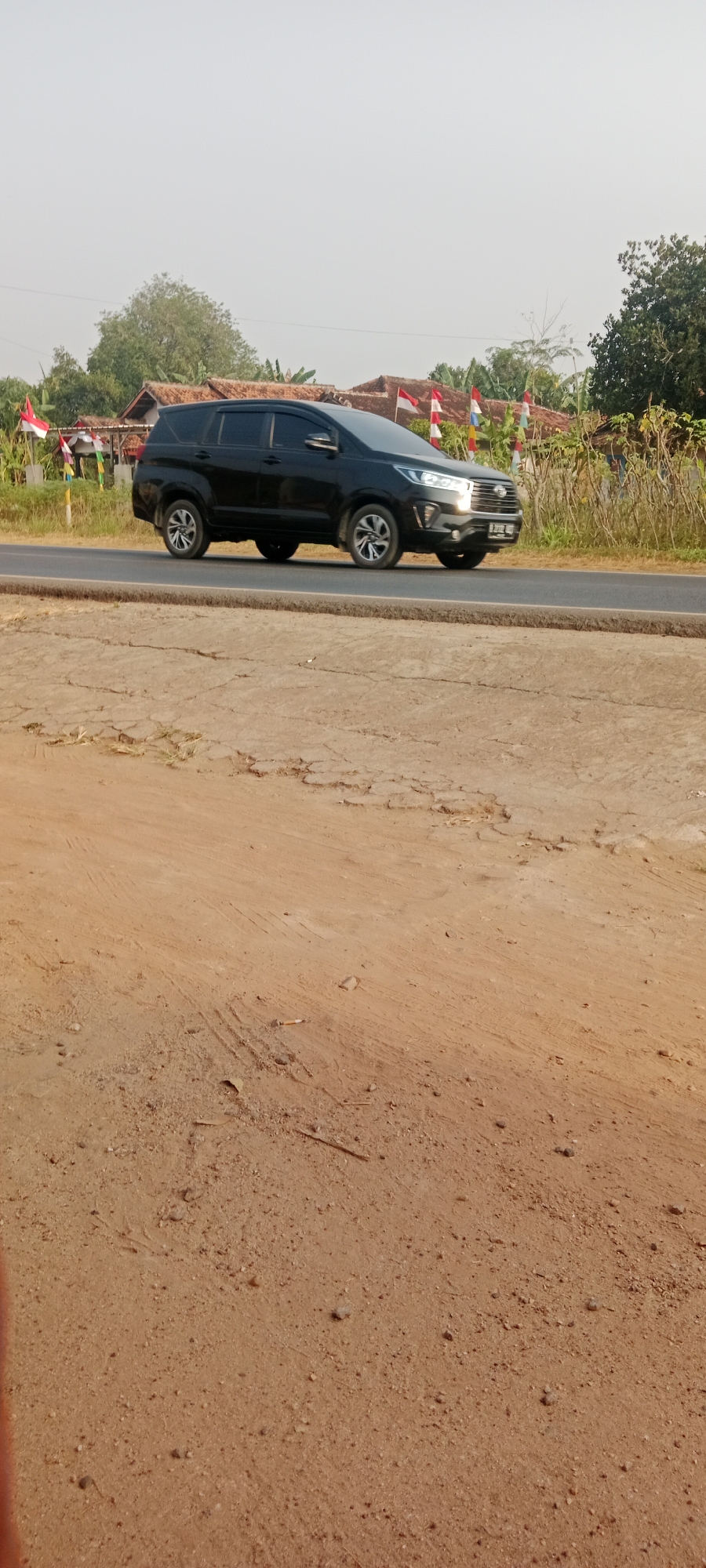 A black Toyota Innova drives on a paved road in Gondang Rejo, Indonesia. The car is captured in profile, moving from left to right. Indonesian flags are visible along the side of the road, suggesting a celebration or national event. The surrounding environment includes houses with tiled roofs and vegetation, indicative of a rural or suburban area. The foreground consists of a dirt road with patches of cracked earth and sparse dry grass. The sky is clear and hazy, suggesting a bright, possibly warm day.