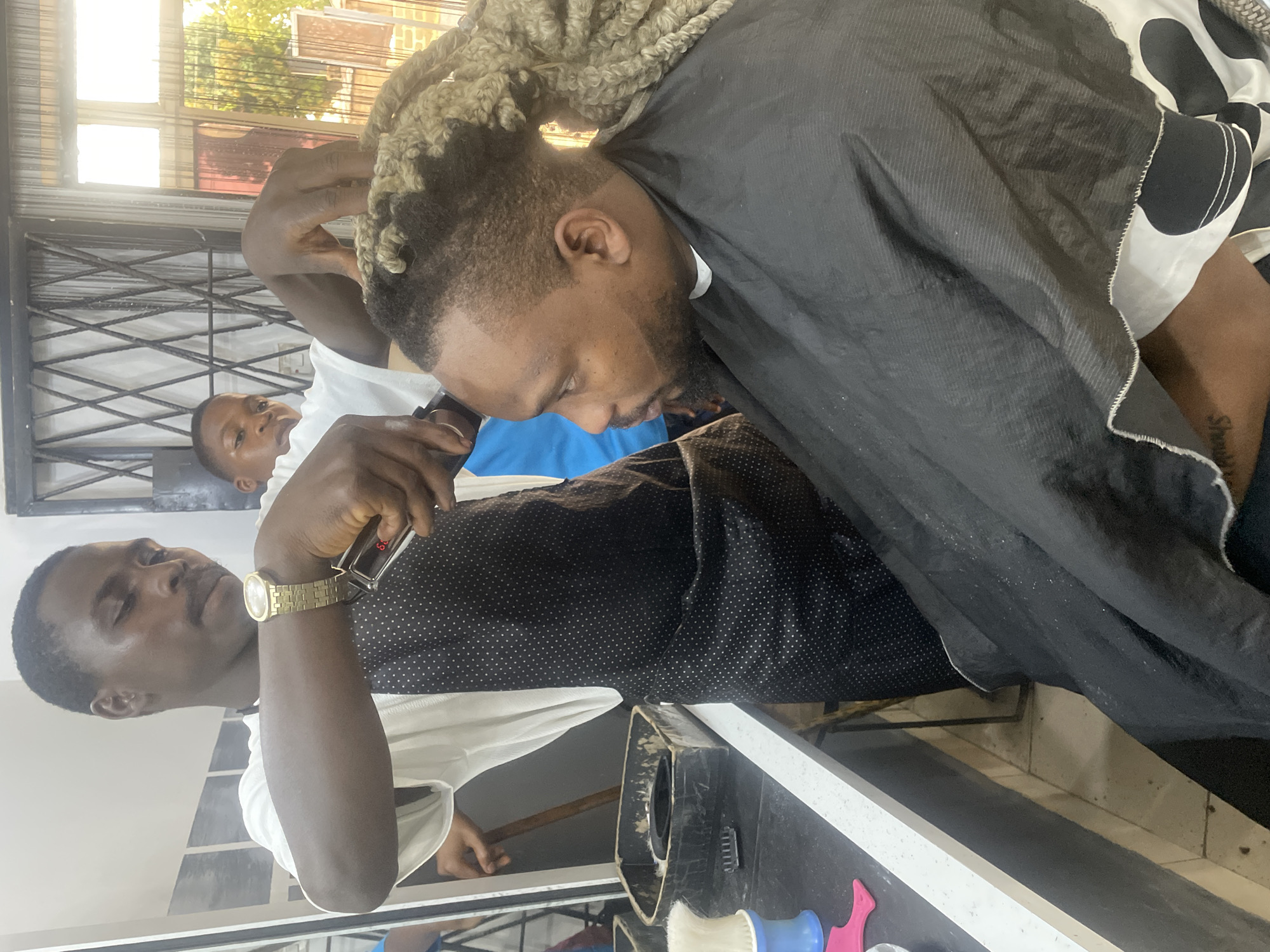 The image shows a man getting a haircut in a barbershop in Jos, Nigeria. The barber, wearing a white shirt and a gold watch, is using clippers on the customer's hair. The customer is wearing a black cape and has blonde dreadlocks. Another man, wearing a white shirt and blue pants, is visible in the background. The barbershop appears to be small and simple, with a window in the background. The image is rotated clockwise.
