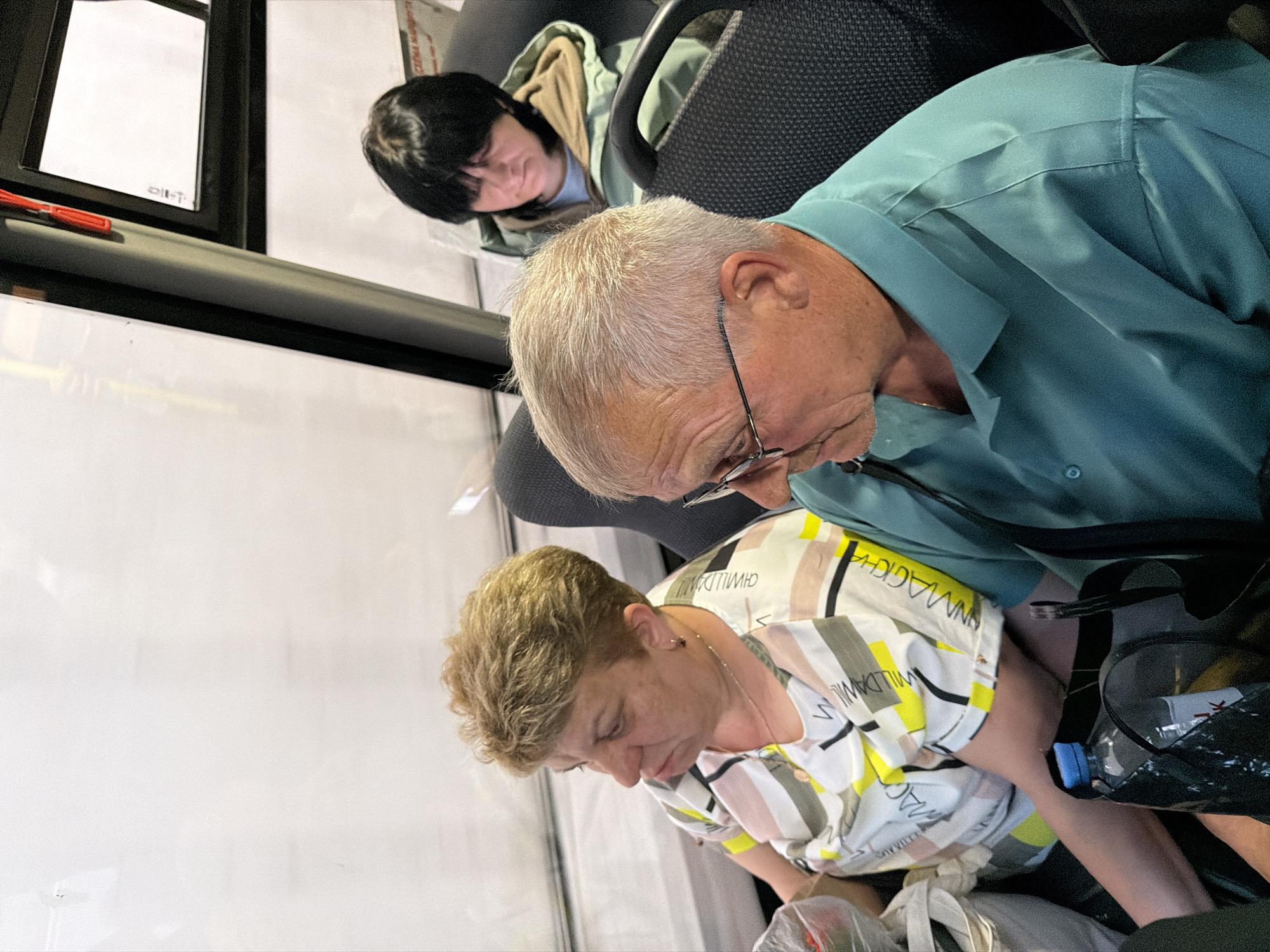 Three individuals are captured in a candid shot. An elderly man with gray hair and glasses is seated in the foreground, wearing a light blue shirt and looking down. A woman with short, blonde hair, wearing a patterned white shirt, is also looking downward, near the man. Further in the background, a woman with dark hair is seated, also appearing to be looking down. The scene appears to be inside a bus or train, as evidenced by the window and seats. The location is Unknown, Unknown.
