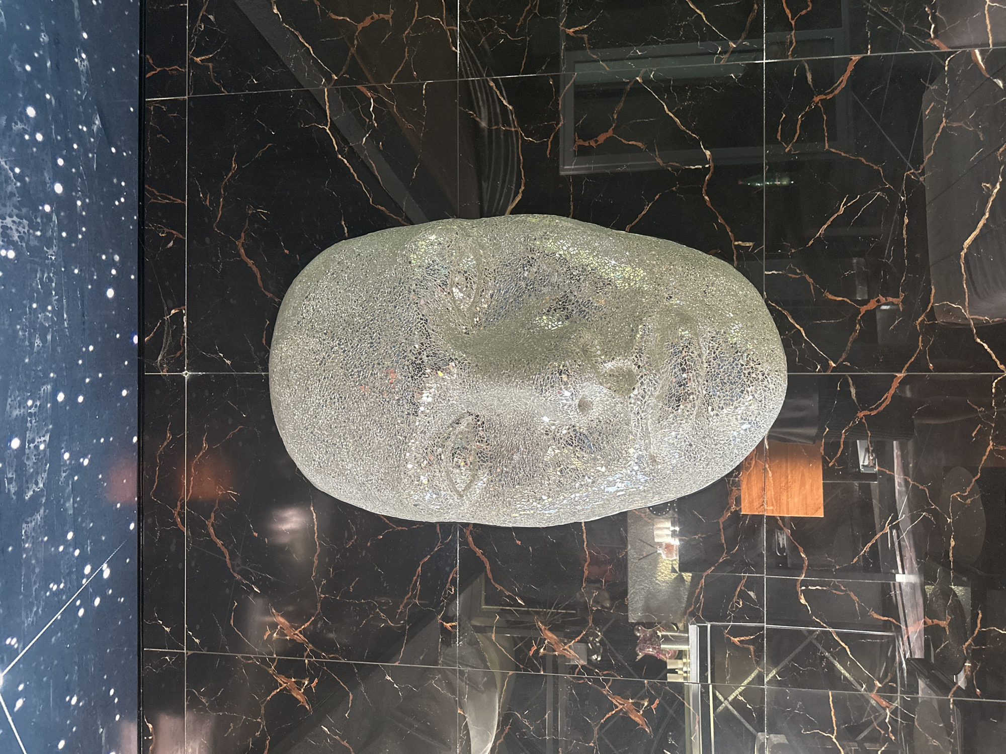 A large, glittery, human-like head sculpture is the central focus of this image, mounted against a wall covered in black marble-patterned tiles with thin, coppery veins. Above the tiled wall is a dark blue ceiling adorned with white speckles resembling stars. The sculpture's surface reflects light, giving it a shimmering appearance. Below the head sculpture, the tiled wall continues, revealing reflections of objects within the room, including what appear to be shelves with bottles and other decorative items, as well as a glimpse of a person's legs and torso wearing dark clothing. The overall environment suggests an indoor, possibly artistic or decorative setting. The image was taken at the hotel Splendid Conference & Spa Resort in Bečići, Budva, Montenegro.