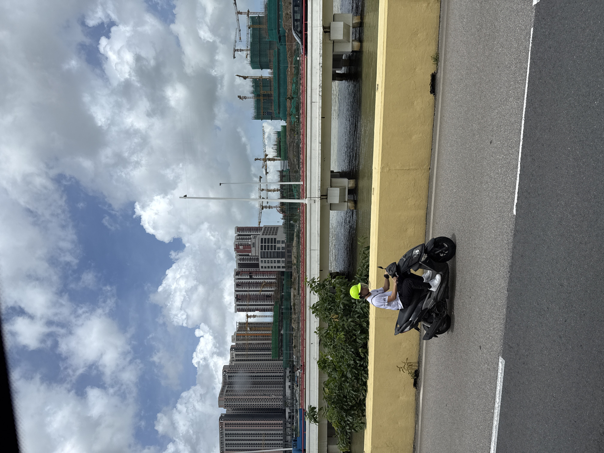 The image captures a street scene in Macau, China, with a focus on a person riding a scooter. The rider, wearing a white t-shirt, dark pants, white sneakers, and a bright green helmet, is positioned on a black scooter. The scooter is on a paved road, with a distinct yellow wall bordering the road on the left. Behind the yellow wall, a body of water is visible, spanned by a white bridge with red pipes running along its sides.
In the background, a cityscape unfolds with numerous tall, modern buildings under construction, characterized by their multi-story structures and the presence of construction cranes. Green netting covers some of the ongoing construction projects. The sky above is partly cloudy, with white clouds against a blue backdrop. The overall atmosphere suggests an urban environment undergoing development.