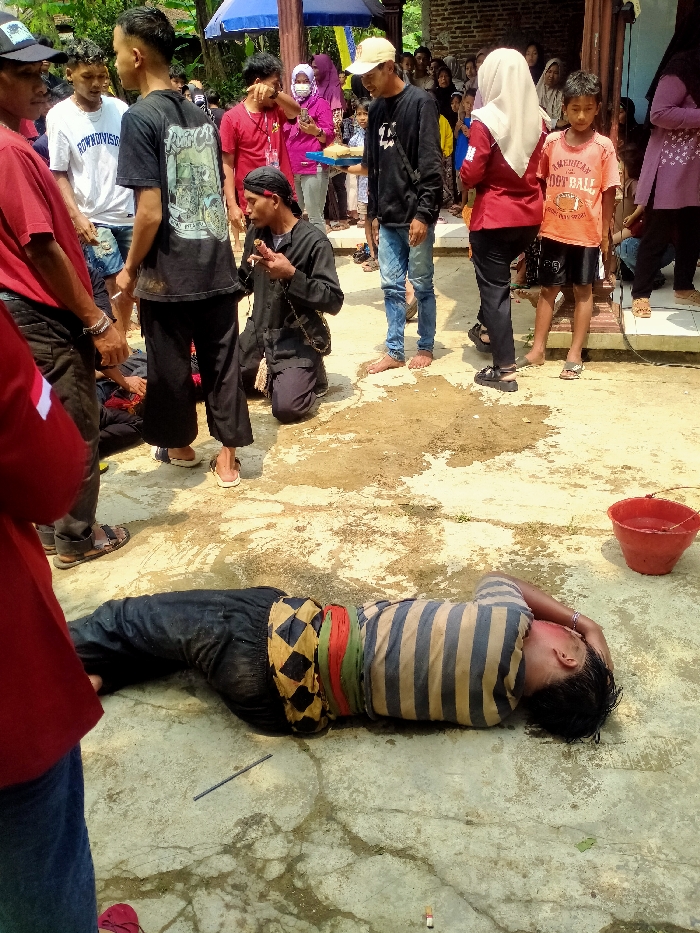 This image depicts an outdoor gathering in Tamansari, Indonesia, taking place on a light-colored concrete surface with dirt patches and moisture stains. The scene is observed by a crowd of approximately 20-30 individuals, many of whom are positioned on a slightly elevated porch area of a brick and wood structure in the background.
In the immediate foreground, a male individual lies prone on his right side on the concrete, facing away from the viewer. He wears black trousers, a multi-colored sash (black and yellow checkered, green, red stripes), and a blue and beige striped short-sleeved shirt. His head is turned to the right, appearing to rest on his left arm, with redness visible on the side of his head/ear. A dark rod-like object lies near his left knee, and a small white cylindrical object is near his feet. To his right, an upright red plastic bucket is positioned on the ground.
To the left of the prone individual, a man in a red short-sleeved shirt and dark trousers stands facing right, partially obscured. Slightly behind him, another man in a black graphic t-shirt and black trousers stands, looking towards the center.
The central focus includes a kneeling man, dressed in a black long-sleeved shirt, black trousers, and a dark headband. A dark chain or strap hangs from his neck. He is kneeling on his left knee, holding a small, light-colored object between his right thumb and forefinger, bringing it towards his mouth as if consuming or presenting it.
Behind the kneeling man, a group of people observe. This includes a man in a white t-shirt, a woman in a purple headscarf and face mask holding a blue tray with yellow items, and a man in a yellow baseball cap, black long-sleeved shirt, and blue jeans, who is barefoot and holds a white rectangular object. To their right, a woman in a maroon top and light beige headscarf stands next to a young boy in an orange "AMERICAN FOOTBALL" t-shirt and black shorts. Both are looking towards the prone individual. The background shows a crowd of spectators, predominantly women in headscarves and children, gathered on the porch of the building. A blue canopy is visible overhead in the background.