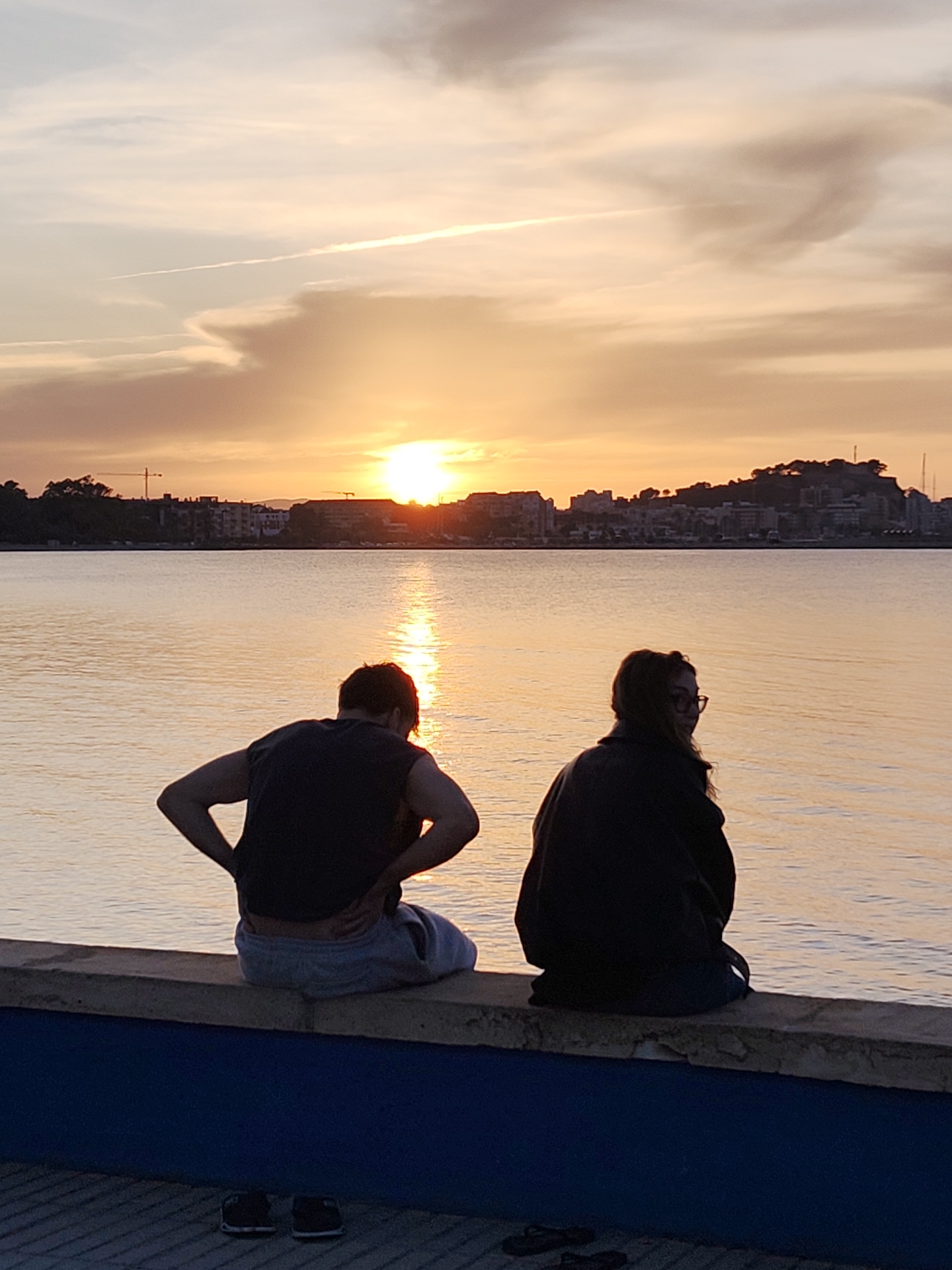 Two individuals are seated on a concrete ledge with a painted blue base, overlooking a body of water reflecting a sunset. To the left is a person with short dark hair, wearing a dark sleeveless shirt and light-colored shorts. Their hands are on their hips. To the right is a person with long dark hair and glasses, wearing a dark jacket. Both are facing the water. The sunset is visible in the distance, over a cityscape with buildings and a small hill. A light contrail stretches across the sky. Pairs of shoes are visible on the ground in front of the ledge. The location is Dénia, Spain, as indicated by the cityscape.
