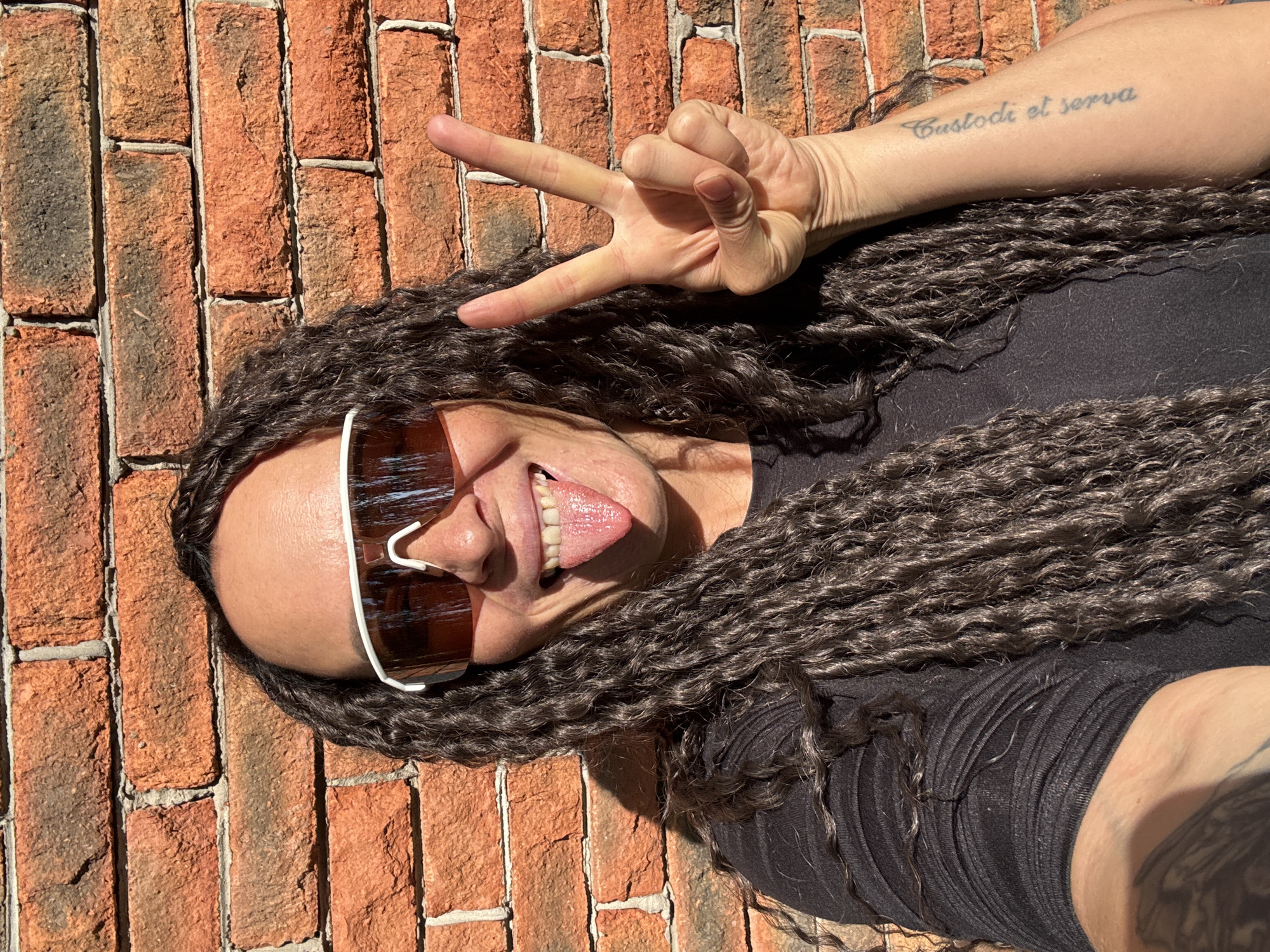 A woman lies on a brick surface, likely outdoors. She is wearing dark sunglasses with a white frame and a black top. Her dark hair is styled in tight braids. She sticks her tongue out and holds up her right hand in a peace sign. A tattoo that reads "Custodi et serva" is visible on her right arm. The image is taken in bright sunlight, casting shadows on the brick surface. There is no indication of the location being Chaikovsky, Russia, unless additional information is available.