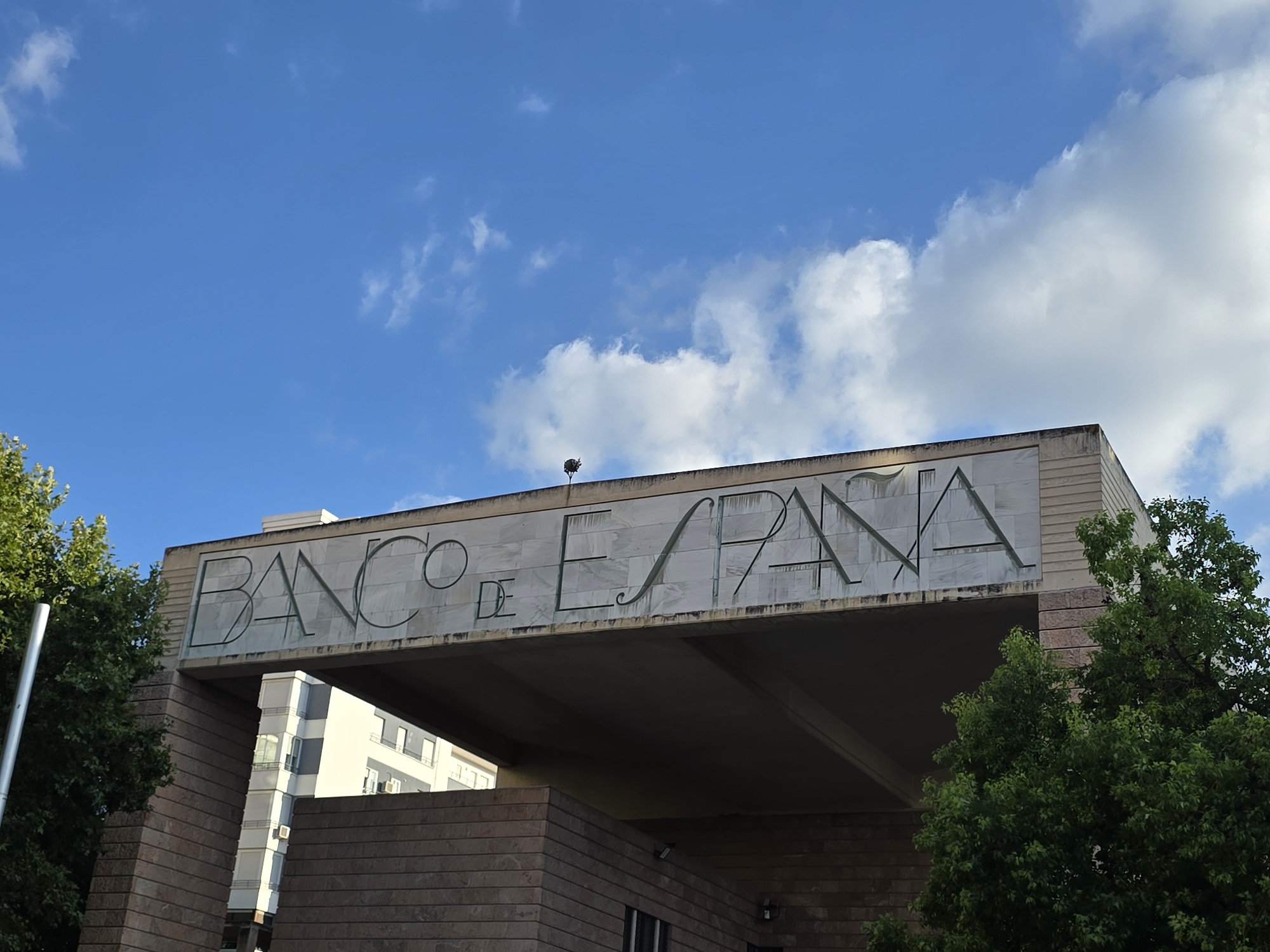 The image shows the exterior of the Banco de España building in Jaén, Spain. The building features a large sign with the words "BANCO DE ESPANA" inscribed in prominent lettering. The architecture appears modern, with stone facade elements and a large concrete overhang. To the left of the building, a tall, thin metal pole is visible, and a lush green tree partially obscures the view of the building's left side. To the right, another dense green tree stands in front of the building. In the background, a multi-story building with white and grey sections and numerous windows can be seen. The sky is a vibrant blue with scattered white clouds.