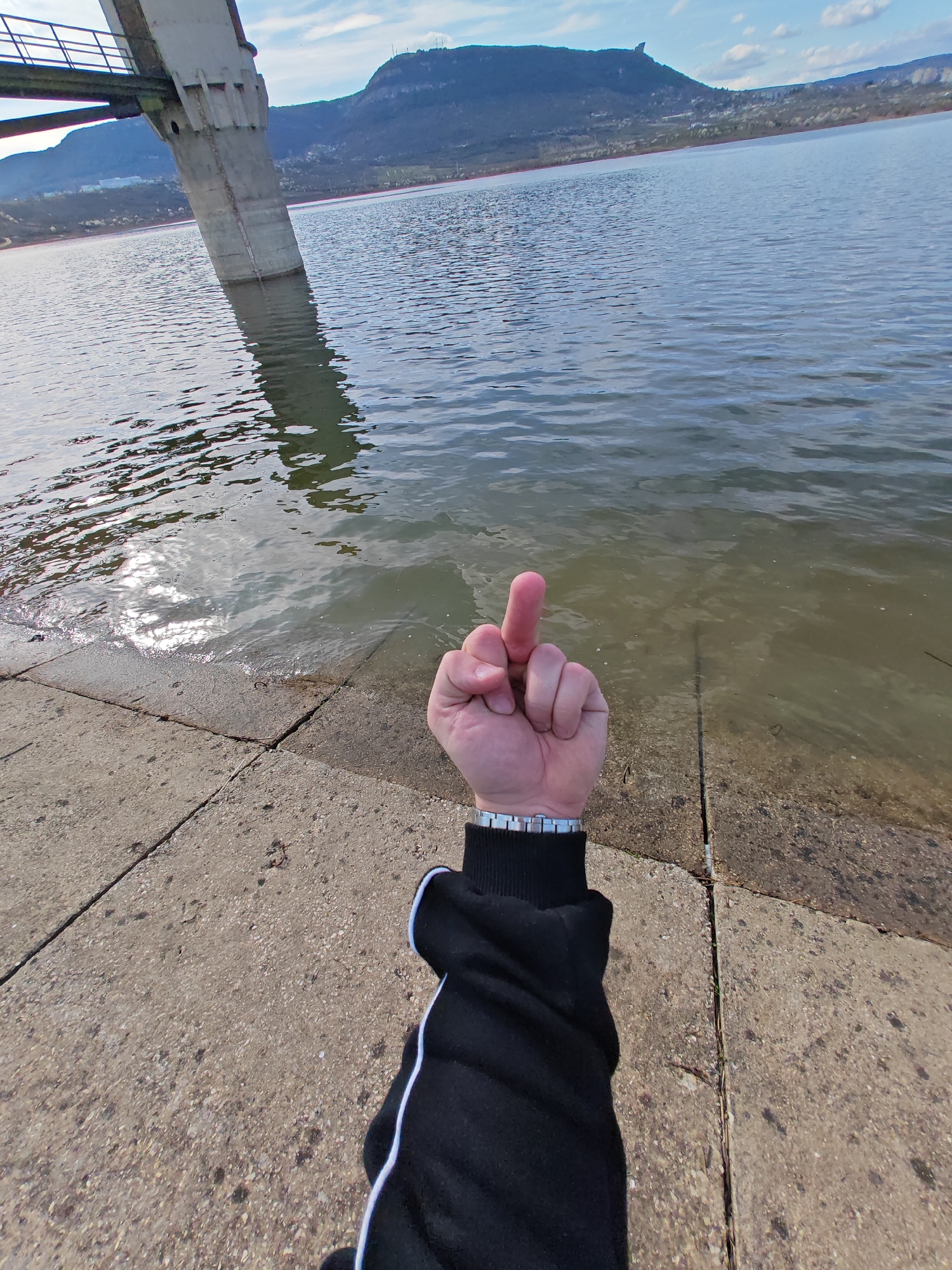 The image shows a person giving the middle finger towards a body of water next to a concrete structure. The location is near a lake, identifiable as the artificial lake "Kyoshkyovete" near Shumen, Bulgaria. In the background, a mountain is visible. The person is wearing a black long-sleeved jacket with a white stripe and a metal watch. They are standing on concrete slabs. The water appears calm.