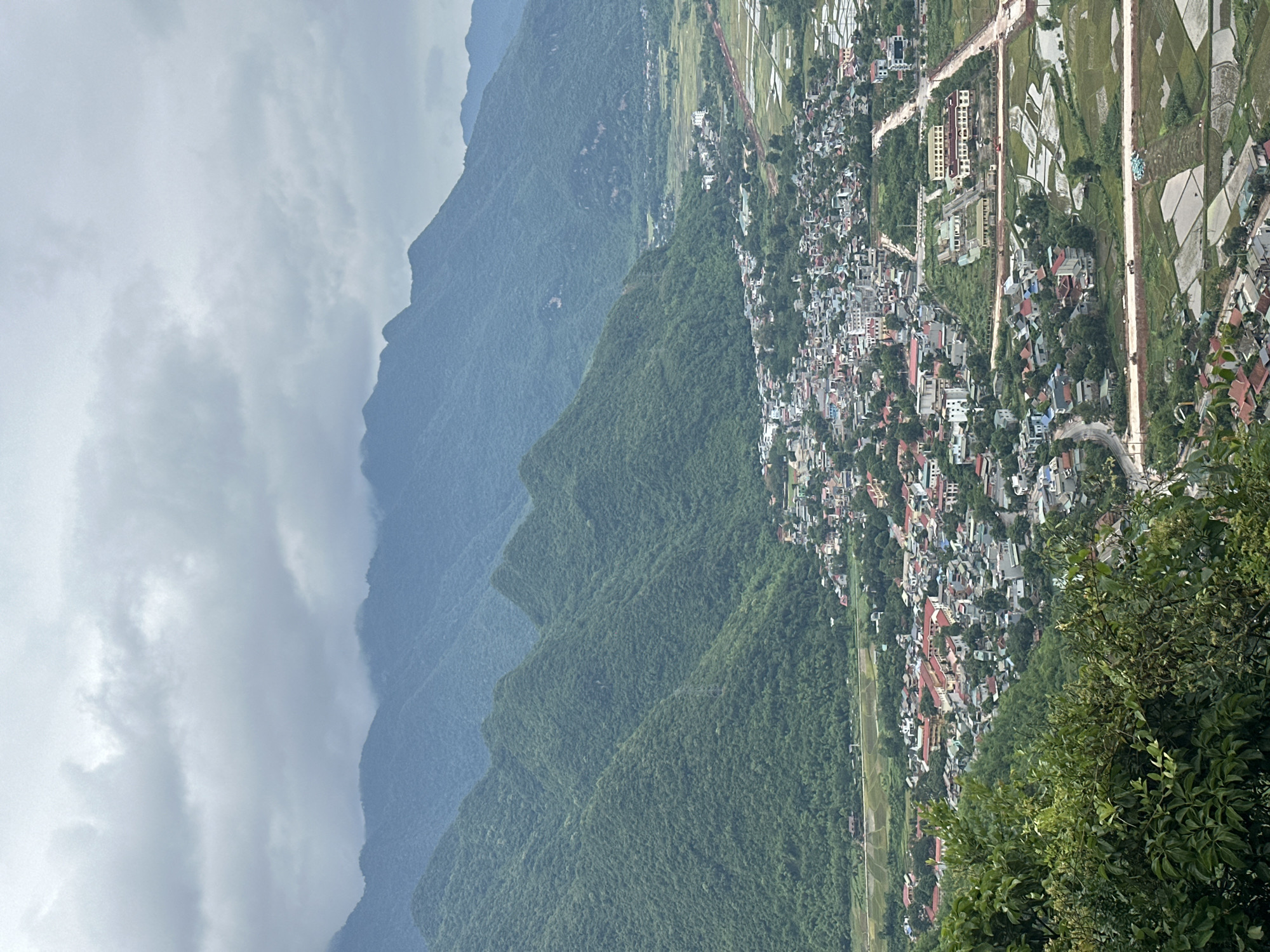 This is a high-angle, wide shot of a valley in Mai Chau, Vietnam, featuring lush green mountains and a developing town. The foreground is dominated by dense green foliage in the lower left corner. A town with numerous buildings, many with red tile roofs, is situated in the valley floor, nestled between the mountains. Roads wind through the town, and construction is visible in several areas, indicated by dirt paths and exposed earth. Surrounding the town are agricultural fields, some appearing to be flooded rice paddies, interspersed with scattered houses and trees. In the background, multiple layers of imposing, densely forested mountains recede into the distance, their peaks shrouded in a hazy, overcast sky. The overall atmosphere is one of natural beauty juxtaposed with human settlement and development. There are no visible people or specific actions depicted in the scene.