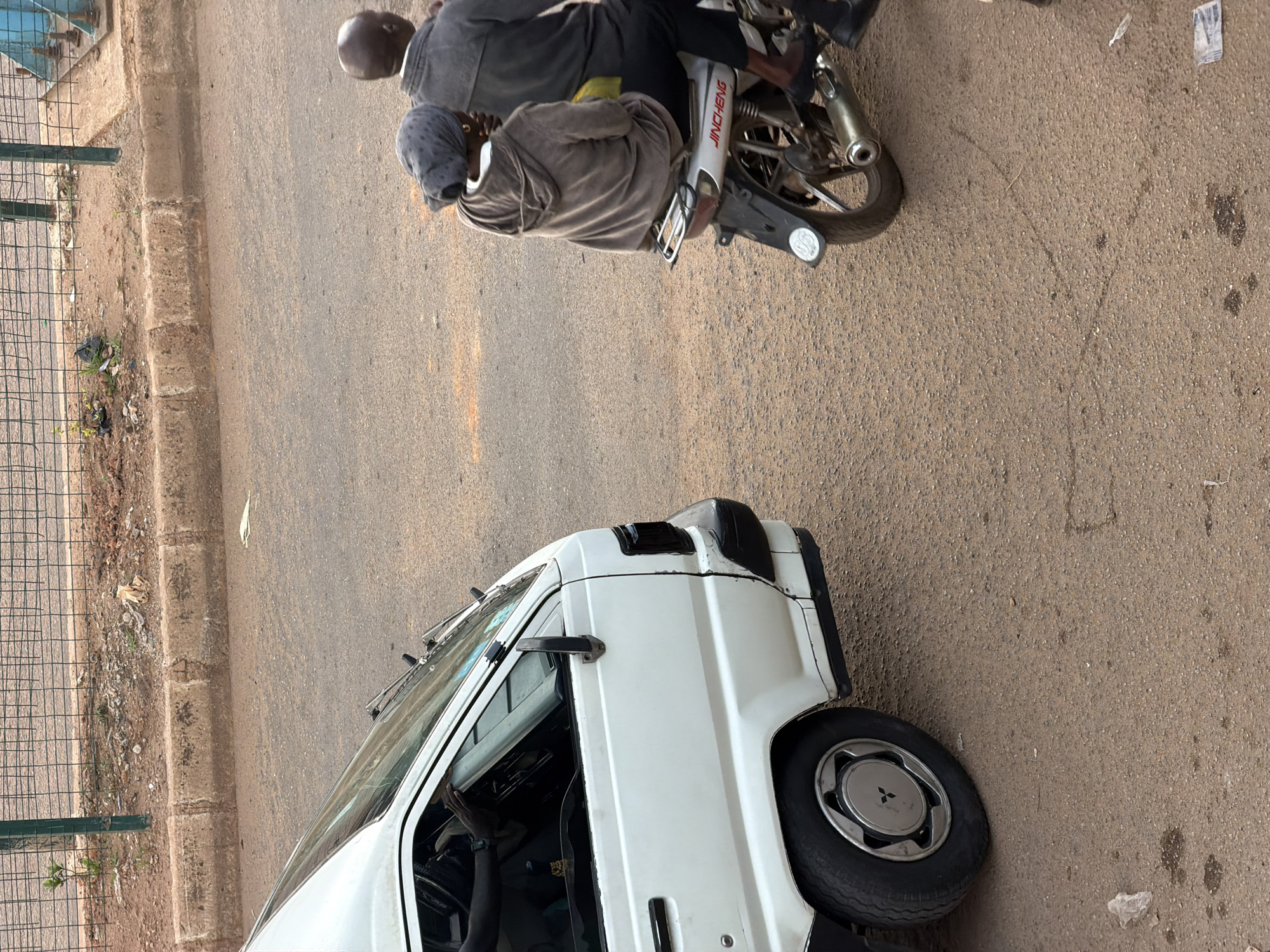 In Bukuru, Nigeria, a white panel van is parked on a dusty, textured road. The van has a black accent near its rear wheel well and appears to be older. To the right of the van, two people are riding a motorcycle, also on the road. The person in front is wearing a grey headscarf and a brown hooded sweatshirt. The person behind them is wearing a grey jacket. The motorcycle is silver with black accents and has "JINCHENG" written on its side. The background shows a curb, a dirt patch with some sparse vegetation, and a metal fence. The overall scene depicts a typical street view in the city.