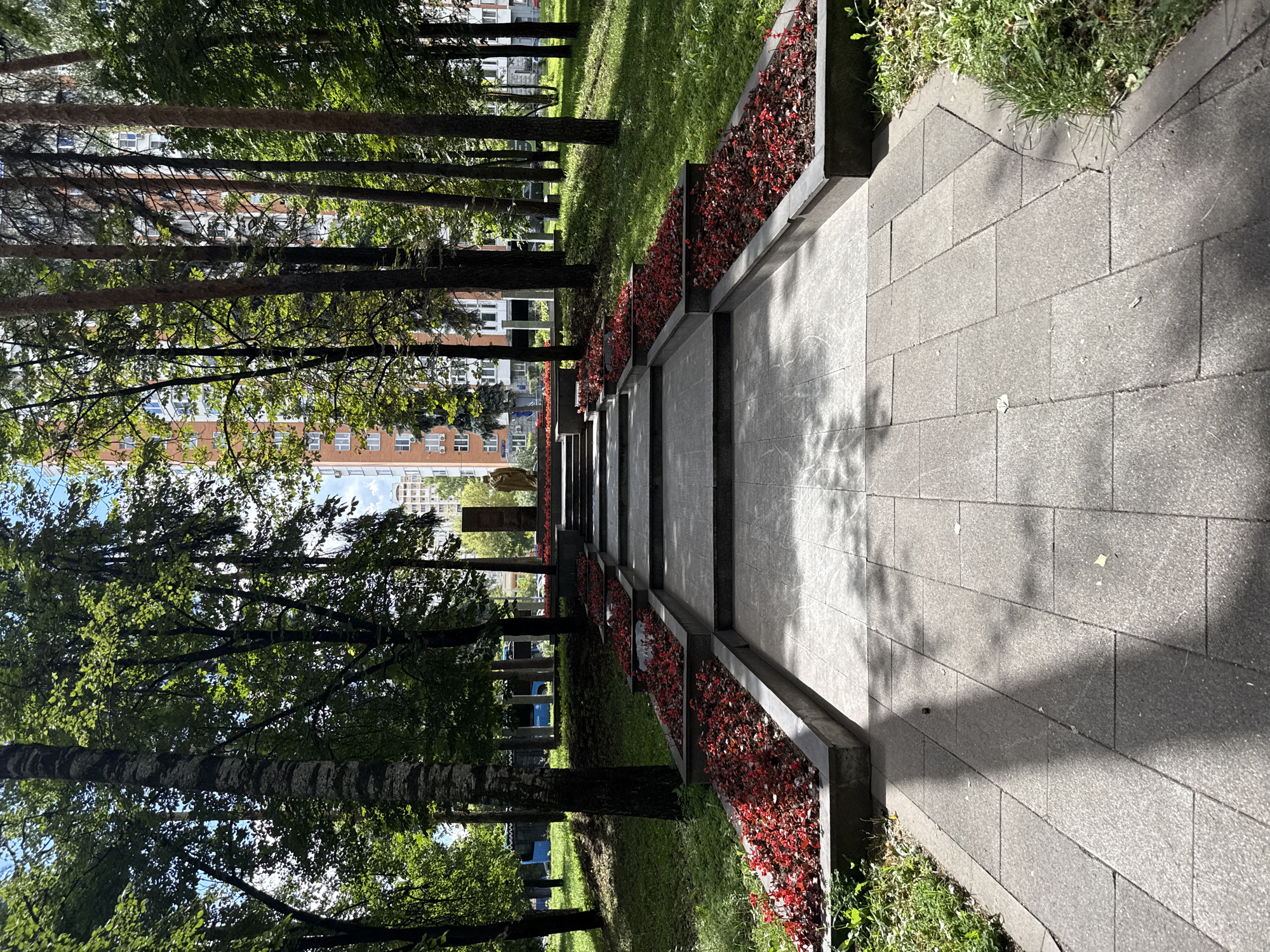 The image depicts an outdoor scene with a pathway leading up a series of tiered steps. The steps are flanked by raised flower beds filled with vibrant red flowers. Tall trees with lush green foliage line the pathway and the surrounding area, casting dappled shadows on the ground. In the background, several tall residential buildings are visible, with one in particular having a distinctive orange-brick facade and numerous windows. At the top of the steps, there appears to be a monument or statue, partially obscured by trees. The sky is clear and blue, suggesting a sunny day. The overall environment is park-like, with well-maintained landscaping. This scene is located in the city of Lyubertsy, Russia.