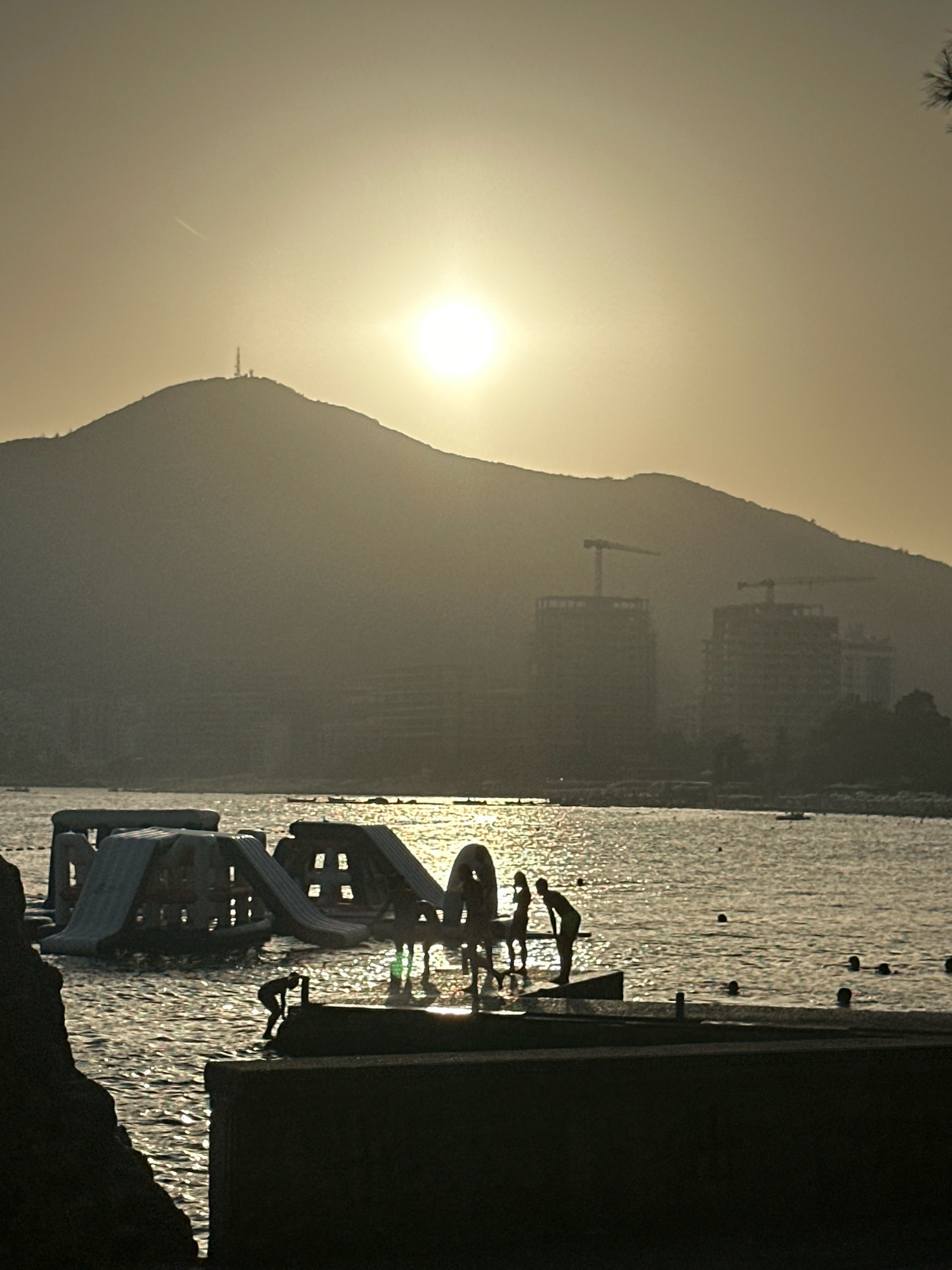 The image depicts a sunset scene at a beach in Budva, Montenegro. The sun, low on the horizon and casting a warm, hazy glow, illuminates the sky and reflects on the rippling water. In the foreground, silhouetted figures are visible on a stone platform by the water's edge, with one individual appearing to be interacting with another or perhaps preparing to enter the water. Further out on the water, a large inflatable water park structure with slides is partially submerged. In the background, a large, tree-covered mountain range rises, with several tall buildings under construction visible at its base, some topped with construction cranes. A radio tower is perched on the peak of the mountain furthest to the left. The overall atmosphere is one of a serene evening by the sea, with elements of both nature and ongoing development.