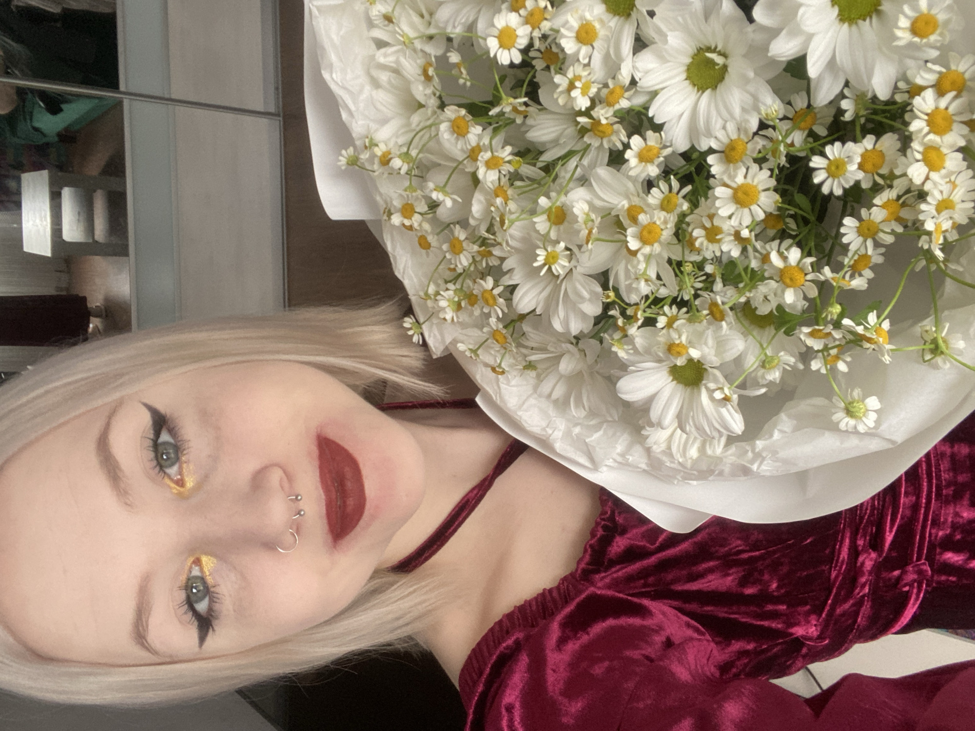 This portrait image captures a young woman with a large bouquet of white daisy-like flowers, presented at an oblique angle. The woman, with blonde hair, light skin, and striking makeup, is positioned with her head tilted significantly to the left, looking directly at the viewer with a subtle smile. Her makeup features dramatic black winged eyeliner, shimmering gold eyeshadow on her inner eyelids, and bold red lipstick. She has a septum piercing (a silver ring) and a small stud or barbell piercing on the side of her nose.
She is wearing a dark red or maroon garment made of velvet, possibly a top or a dress, which features thin straps. The bouquet, occupying the upper right portion of the frame, is densely packed with numerous white daisy-like flowers, characterized by their white petals and distinct yellow or green-yellow centers. The flowers are wrapped in white paper.
The background behind the woman's head and upper body is a reflective surface, likely a mirror, showing blurred reflections of interior elements such as shelves and indistinct objects. The overall setting appears to be indoors. No specific time of day or weather cues are visible, but the indoor lighting suggests it could be any time. There is no visible text in the image. The location context for this scene is Izhevsk, Russia.