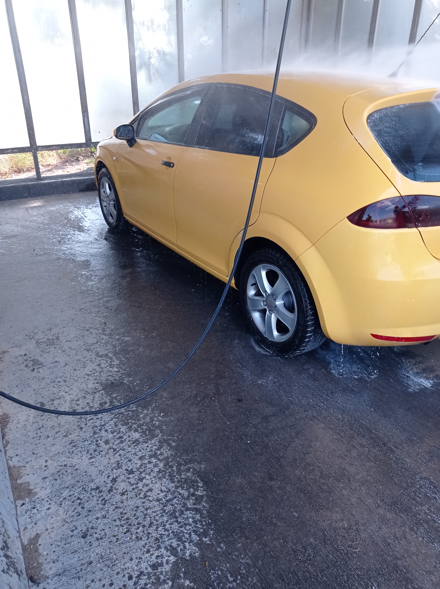 The image shows a yellow car being washed at an outdoor self-service car wash. The car is a hatchback. A black hose from a pressure washer is visible, spraying water onto the car, particularly the roof and rear quarter panel. The ground is wet and reflective, indicating water and soap suds. The car wash bay has translucent, ribbed panels forming a partial enclosure, allowing daylight to filter through. The background shows some dry vegetation and the sky. The location is the self-service car wash at the Shell petrol station on the M43 motorway, just outside Szeged, Hungary, near the Hungarian-Serbian border. No people are visible in the frame.