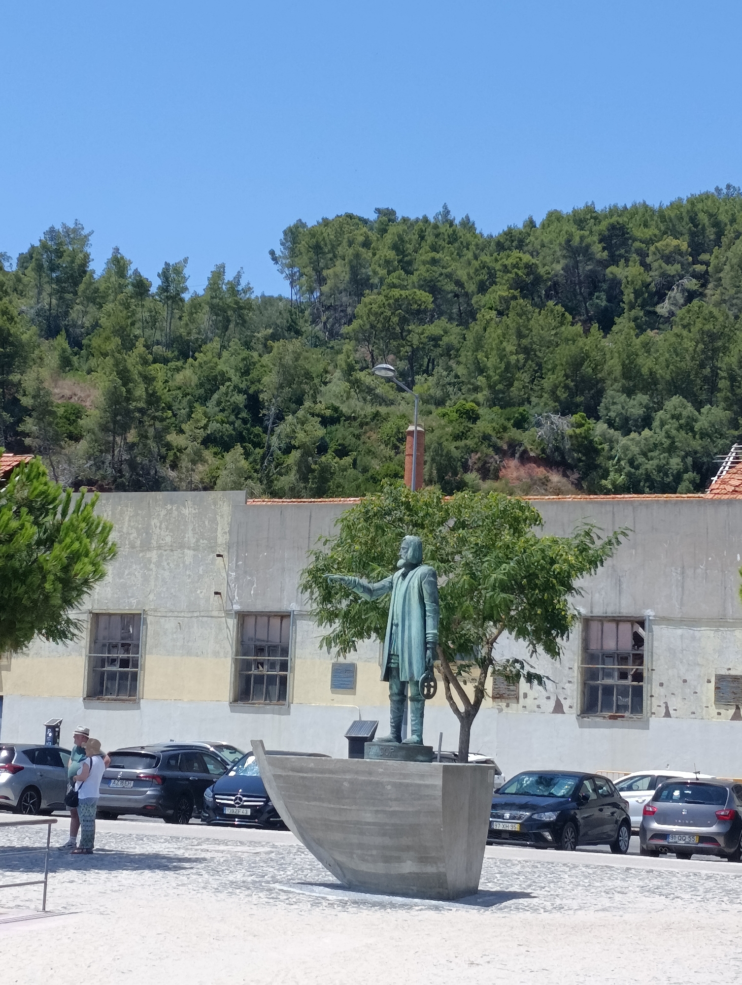 A full shot shows a statue of a man in historical clothing standing in what appears to be a public square in Setúbal, Portugal. The statue is positioned in front of a weathered building with large, broken windows, and a lush green hillside covered in trees rises behind it. In the foreground, a large boat-like structure, possibly part of the statue's base or a separate art installation, is visible. Several cars are parked in the background, suggesting a busy area. Two people, a man and a woman, are standing near the cars, appearing to be observing the statue or the surrounding area. The sky is a clear, bright blue. The overall impression is of a sunny day in an urban setting with a historical monument as the focal point.