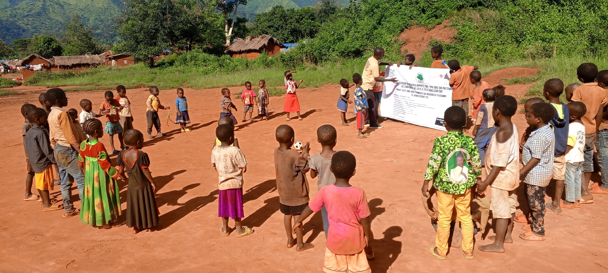 In Ngalula, Democratic Republic of the Congo, a brightly lit outdoor scene depicts approximately 30-40 individuals, primarily children, gathered on an expansive reddish-brown dirt ground. Two adult males stand centrally on the right, holding a large white rectangular banner horizontally. The banner displays text, logos including a green tree and a red cross-like symbol, and mentions "TPC / DRC UN PARTENAI," "DE COMPAGNIE 2026 A 2076 BAILLEUR DOC," and other details in French. A large group of children, varying in age, are positioned in a loose semi-circle around the banner and the adults. Many children are barefoot or wear sandals, dressed in varied, well-worn clothing. A small black and white soccer ball lies on the ground to the left of the banner. In the background, lush green foliage, trees, and a verdant hill are visible under a clear sky. Several single-story, rustic structures with brown roofs and reddish-brown walls are located in the mid-ground on the left, partially obscured by greenery. The bright sunlight casts distinct shadows on the ground, indicating a clear day. The children appear to be observing the banner and the adults, suggesting a presentation or gathering.