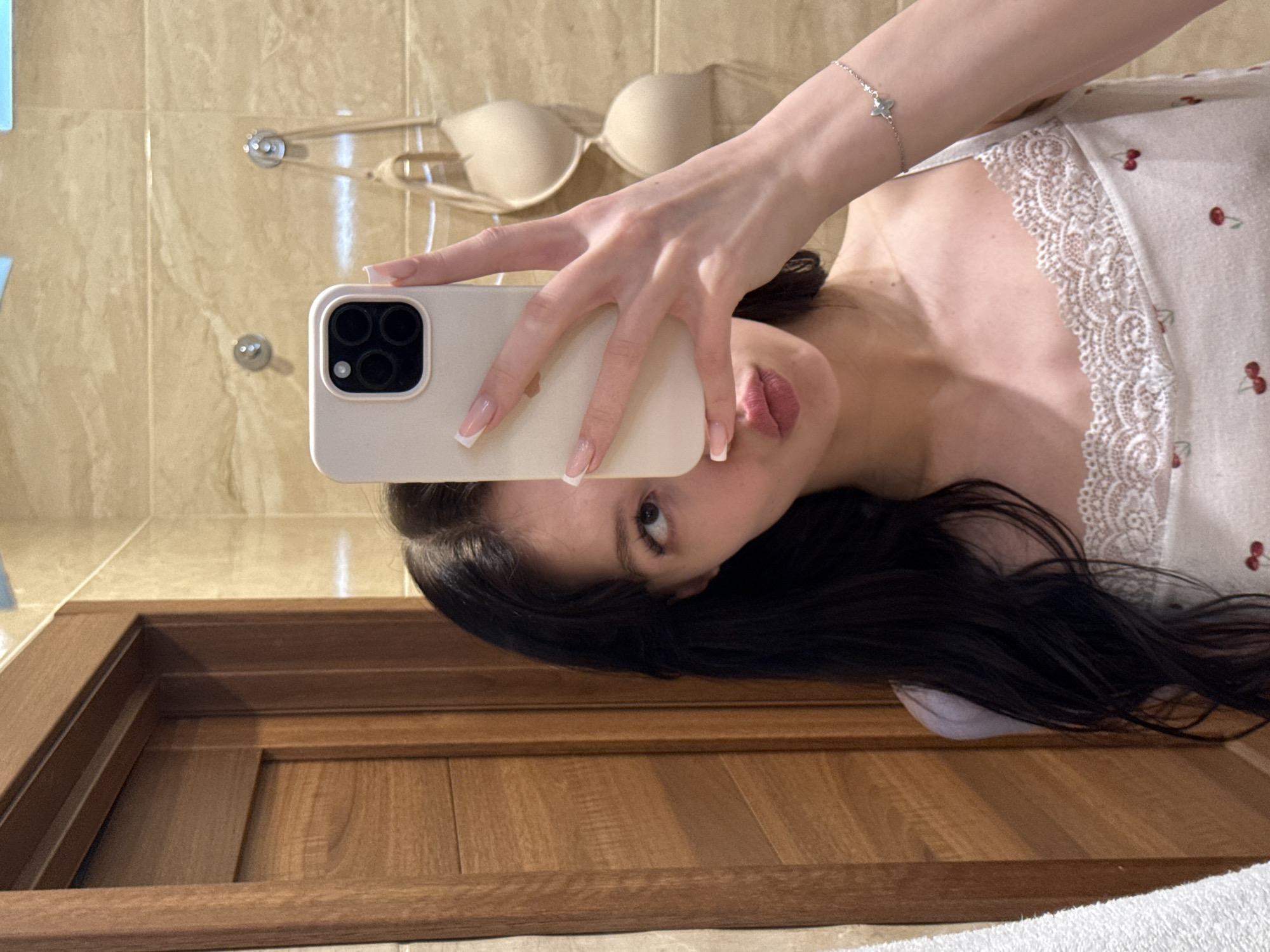 A young woman with long, dark hair is taking a mirror selfie in what appears to be a bathroom in Khimki, Russia. She is wearing a white camisole with a cherry print and lace trim. She is holding up a light-colored smartphone, obscuring the lower half of her face. Behind her, two beige bras are hanging on hooks against a tiled wall. To her left is a wooden door.