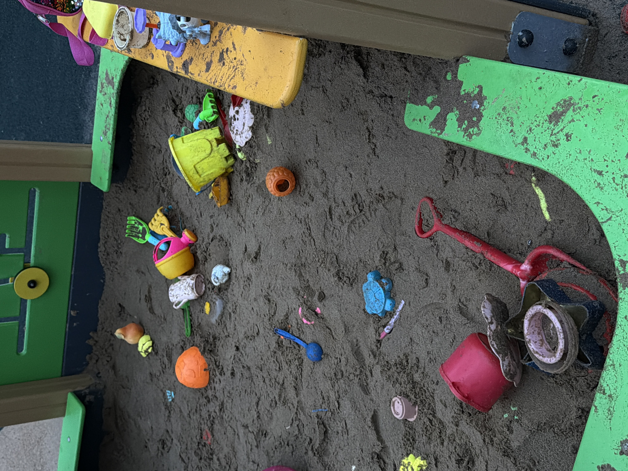 The image depicts a close-up view of a sandbox filled with dark, granular sand. The sandbox contains a variety of scattered children's toys, including buckets, shovels, a watering can, various plastic molds shaped like animals and objects, and a red plastic rake. Some of the toys are partially buried in the sand. The sandbox appears to be part of an outdoor playground structure, with glimpses of green and yellow plastic components of the structure visible at the edges of the frame. The overall scene suggests a recently used play area. There is no indication of the location in the city of Фролы, Russia, within the image.