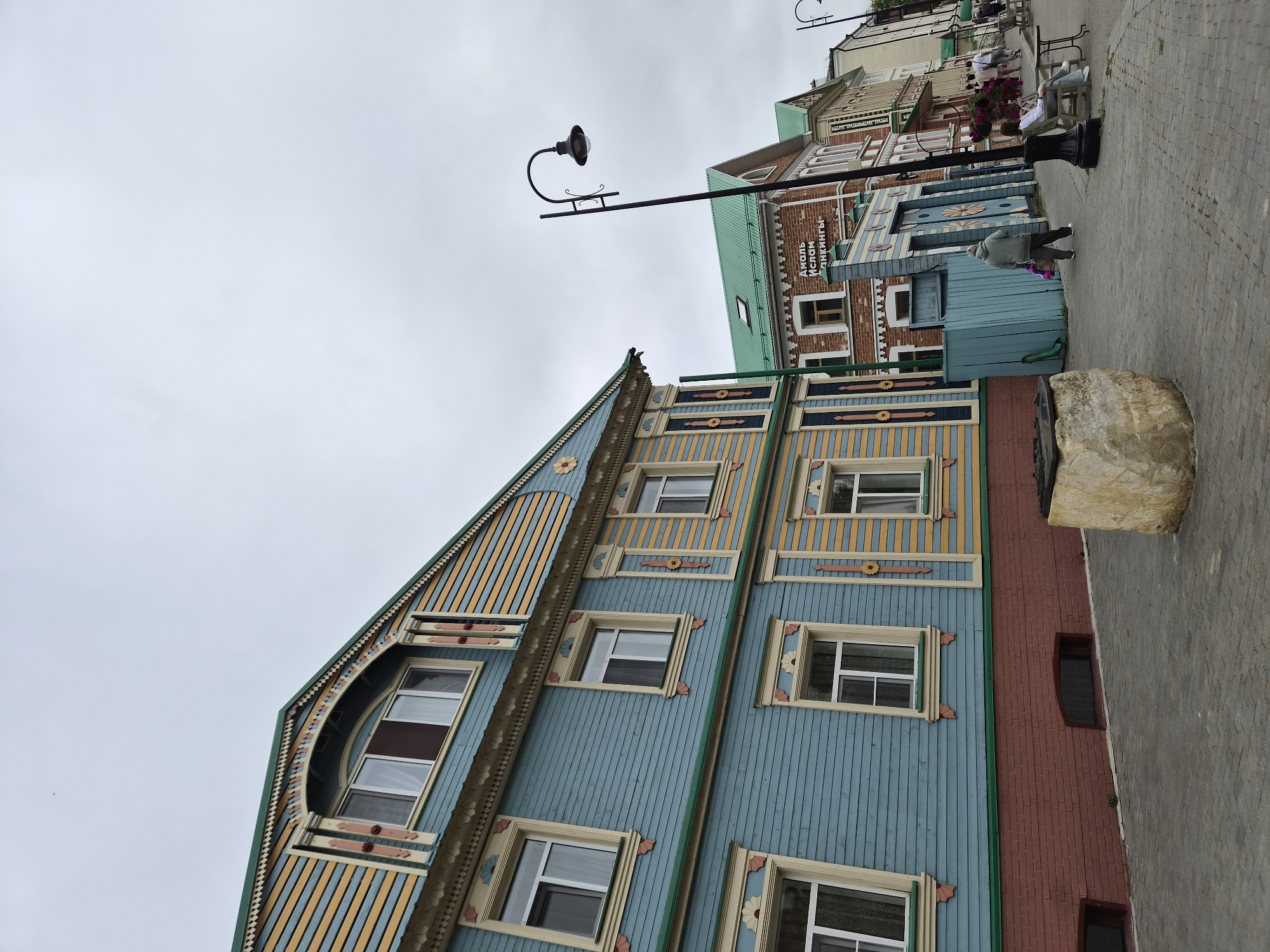 The image depicts a street scene in Kazan, Russia, characterized by traditional Tatar architecture and a cloudy sky. On the left, a brightly colored wooden house features intricate carvings and ornamentation in yellow, blue, and red. The house has multiple windows with decorative frames. In front of the house, a stone structure, possibly an old well, sits on a paved walkway.
Further down the street, other buildings display similar architectural styles, with some having brick facades and green roofs. One building has text in Cyrillic script, which reads "Амаль Ислам Янгинты" (Amal Islam Yangynty), suggesting a commercial establishment.
Several people are present in the scene. A person in a grey hoodie walks away from the camera carrying shopping bags. Further along the street, a woman sits on a bench next to a table, and a group of people are walking away from the camera. A black lamppost with an ornate design stands on the right side of the walkway. The pavement is made of gray interlocking bricks. The overall atmosphere is overcast, indicating a cloudy day.