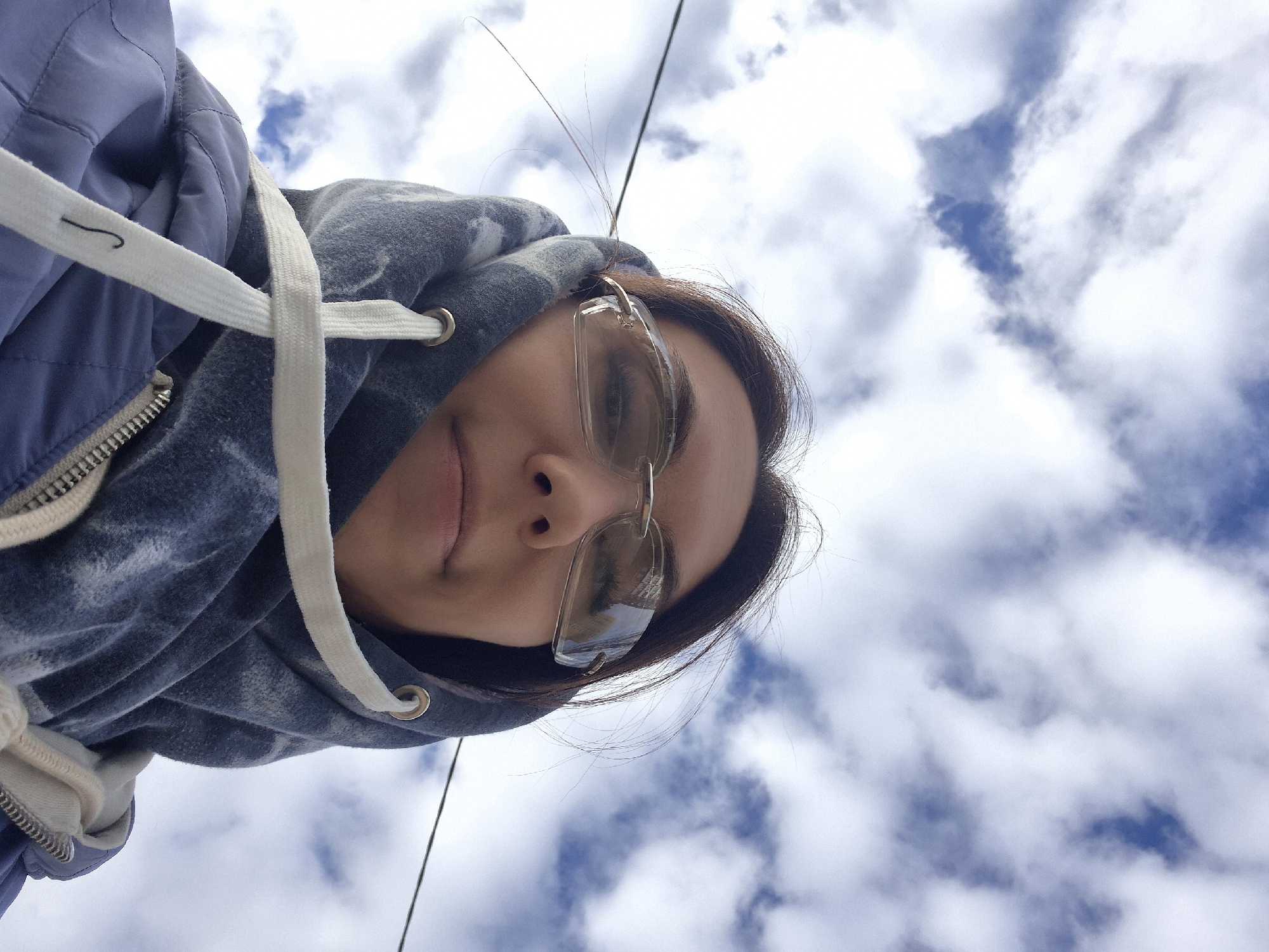 A low-angle, waist-up, medium shot captures a young woman outdoors, looking directly at the camera. She is wearing clear, rimless glasses and a blue hooded jacket, with the hood pulled up and tied with white drawstrings. Her hair is dark brown and loose around her face. The background is a sky filled with white and gray clouds, with glimpses of blue sky. A thin black wire, possibly a power line, is visible across the frame. The scene appears to be set in Chaikovsky, Russia, as indicated by the presence of a building reflected in her glasses. The overall impression is a casual, self-taken photograph.