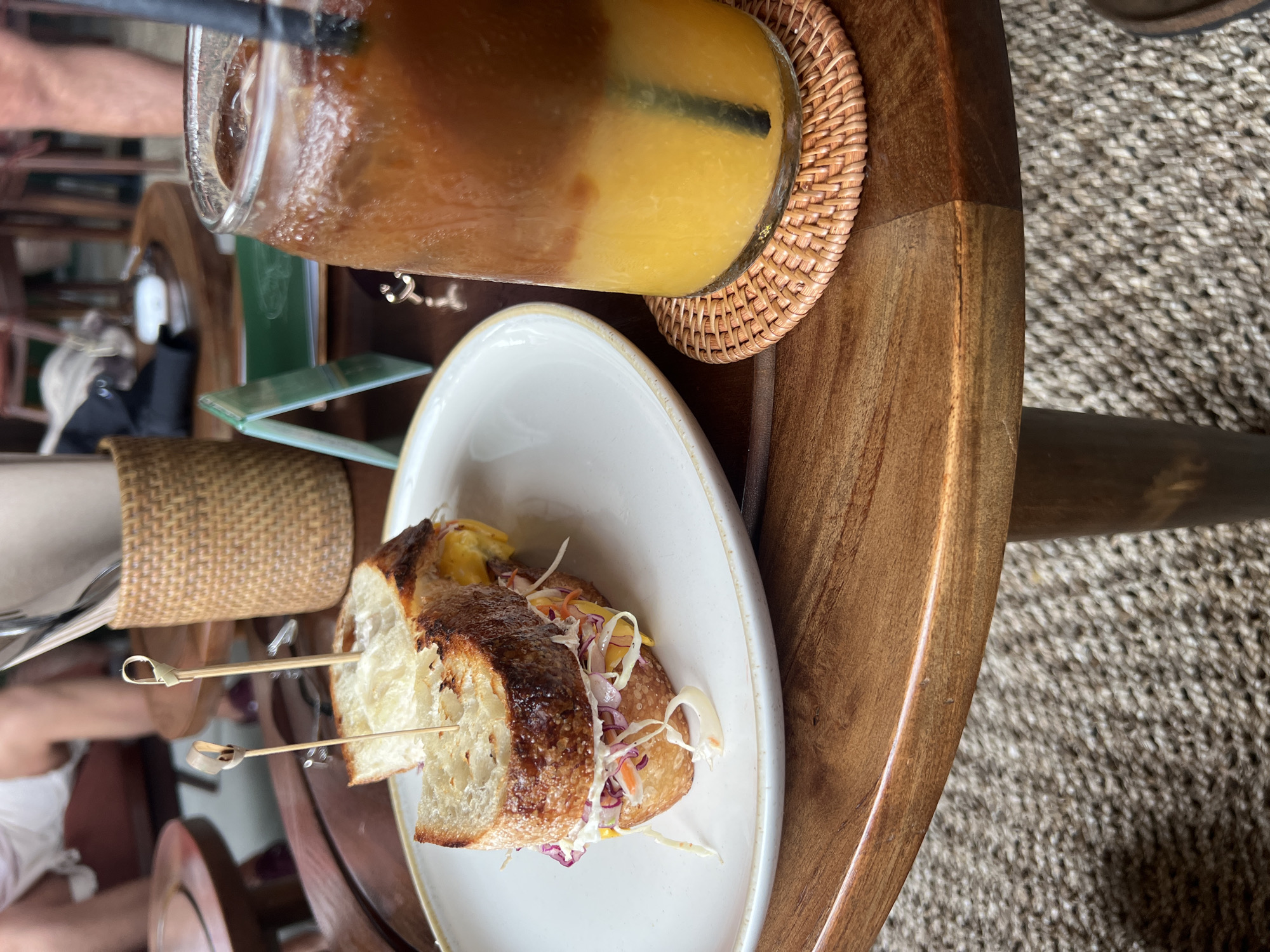 A high-angle shot captures a casual dining setting in North Kuta, Indonesia. On a light-toned wooden table, a clear glass contains a layered beverage with a dark top layer and a lighter yellow/orange bottom layer, suggesting a juice blend. A black straw is inserted into the drink, and the glass rests on a round, woven rattan coaster.
Adjacent to the drink, a white, irregularly shaped plate holds a sandwich or baguette, cut into two halves. The bread has a crispy, golden-brown crust. The filling consists of a yellow ingredient, possibly cheese or a sauce, along with a colorful slaw-like mixture of shredded red cabbage, white cabbage, and possibly carrots. Two decorative wooden skewers with loop handles are used to hold the sandwich halves together.
In the blurred background, a woven basket-like object and a green item, possibly a menu, are partially visible. The glossy wooden table surface shows faint reflections of the surroundings. The floor beneath the table is covered by a coarse, light-colored woven mat. The natural lighting indicates a daytime setting, suggesting a relaxed meal or snack. No visible text is present in the image.
