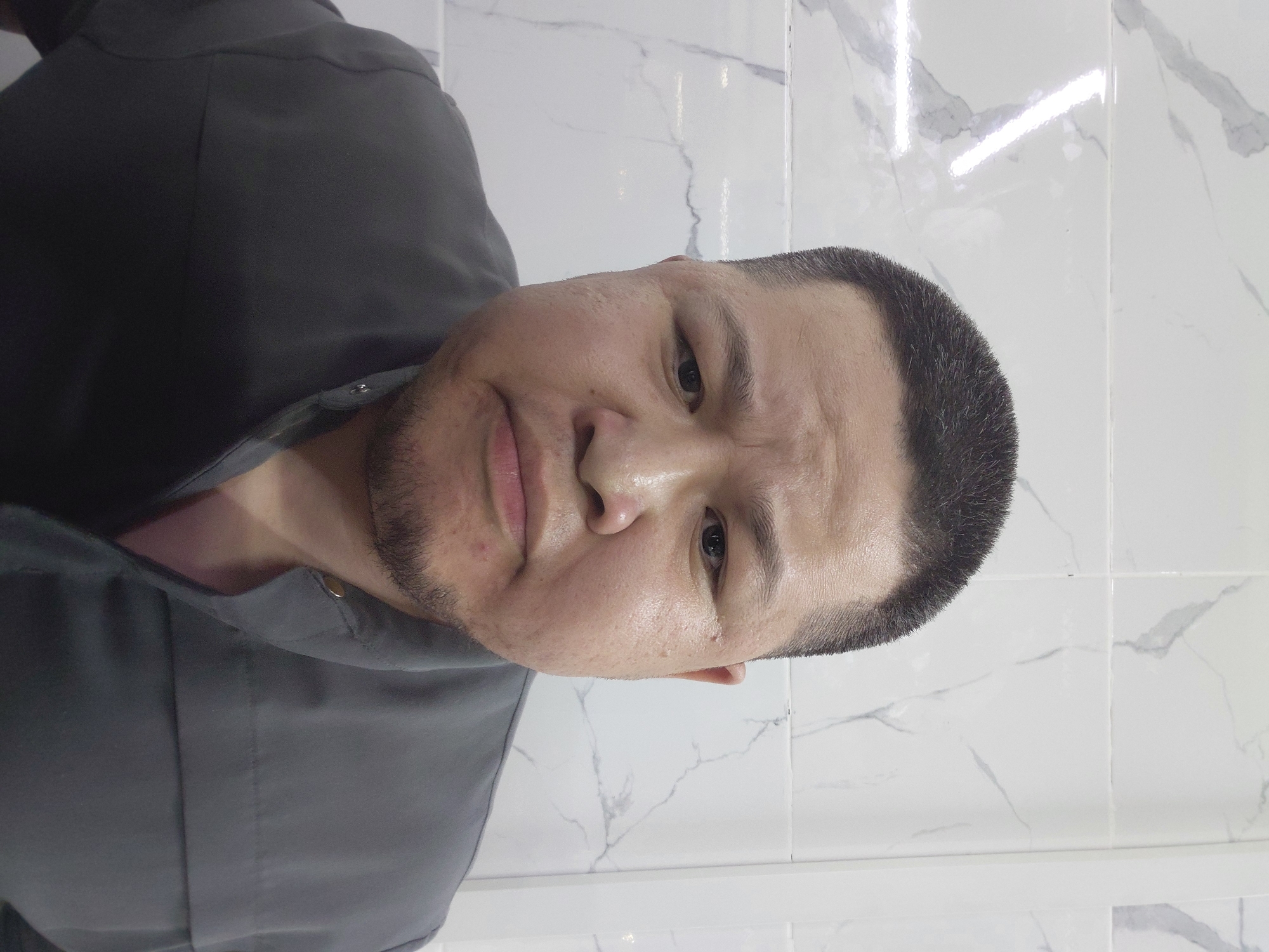 A medium shot shows a man with a buzz cut and light stubble, wearing a dark gray collared shirt. He is looking directly at the camera with a neutral expression. The background consists of a white tiled wall with gray veining and a vertical white trim piece on the right side. The lighting appears to be overhead, creating a slight glare on the tiled surface. The image appears to have been taken indoors, possibly in a bathroom or a similar tiled environment, and its metadata indicates it was taken in Aktau, Kazakhstan.