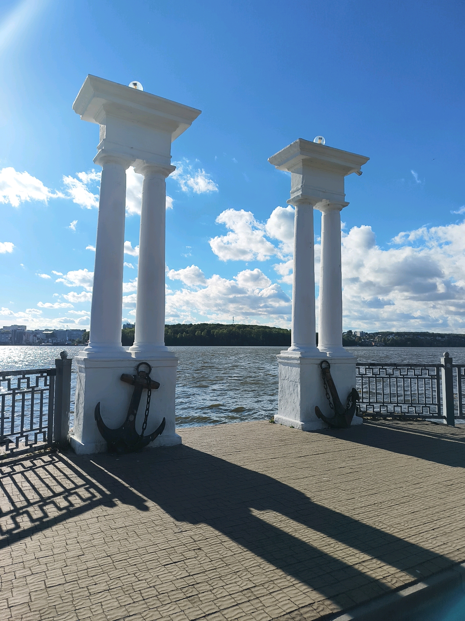 The image depicts a waterfront promenade with two white classical-style pillars, each topped with a small decorative light fixture. An anchor is attached to the base of each pillar. The pillars stand on a paved walkway, which casts long shadows from the afternoon sun. To the left and right of the pillars, a black metal railing lines the edge of the promenade. Beyond the railing, a body of water, likely a lake or river, is visible. In the distance, a treeline separates the water from a cityscape featuring buildings. The sky is bright blue with scattered white clouds. This scene is located at the Ternopil Lake embankment in Ternopil, Ukraine.