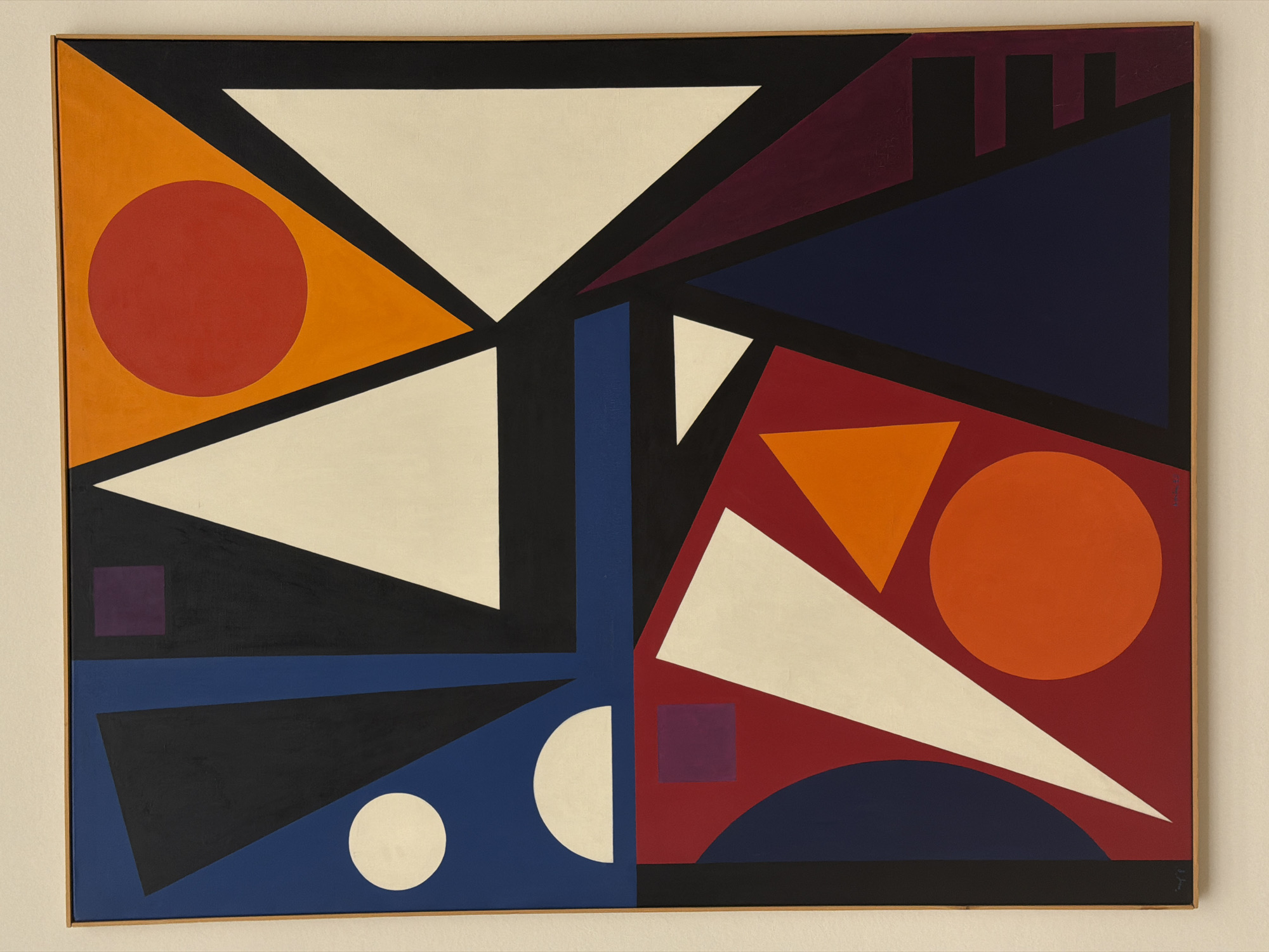 This is a forensic, matter-of-fact description of an abstract geometric painting. The painting is framed in light wood and mounted on a neutral-colored wall, suggesting it is on display in a gallery or museum. The artwork features a vibrant composition of interlocking geometric shapes in bold colors, including red, orange, blue, black, white, and purple. There are triangles, circles, squares, and semi-circles arranged in a dynamic and balanced manner. The image does not depict any people, actions, or interactions. The setting is a sterile exhibition space, typical of a museum in Basel, Switzerland, such as the Kunstmuseum Basel, which houses a significant collection of modern and contemporary art.