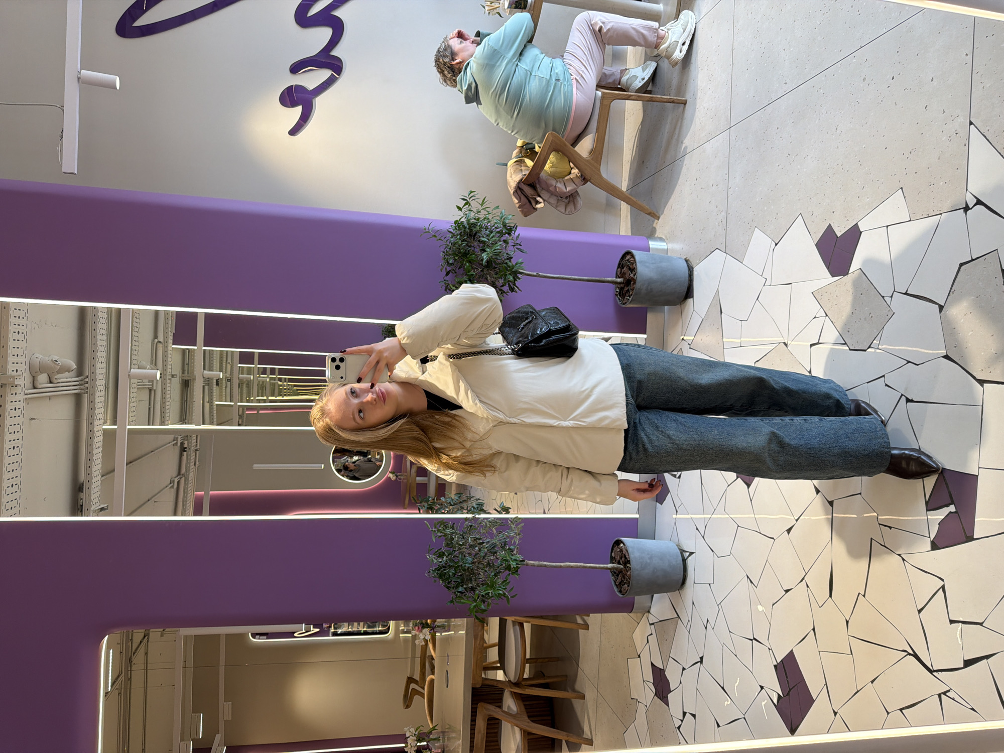 A woman stands in front of a mirror holding a mobile phone, with a purple rectangular border reflecting the surrounding environment. The scene appears to be indoors, possibly a salon or cafe, as suggested by the purple design elements and the presence of seating and small potted trees. The woman in the mirror is wearing a light-colored jacket, jeans, and dark shoes, with a small black bag hanging from her shoulder. In the reflection of the mirror, there's another person sitting on a chair wearing a light blue hoodie and light pink pants. The floor has a distinct mosaic pattern with white, gray, and purple tiles. The reflection also reveals part of the interior architecture, including white shelving and lighting fixtures. Based on the information provided, this image was taken in Krasnodar, Russia.