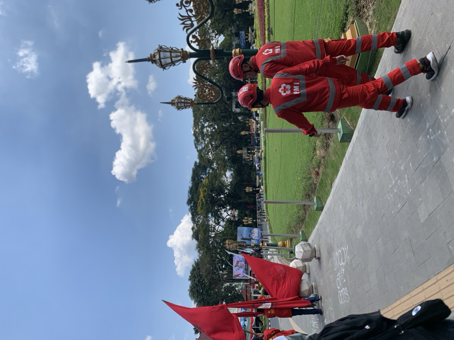 Two men wearing red uniforms with the logo "PMI" are walking on a paved sidewalk in front of a grassy area. The sidewalk has white chalk markings and a street lamp with a golden top. In the background there are trees, a red flag on a pole, and a blue sky with white clouds. The scene is in Malang, Indonesia, at the coordinates -7.977 latitude and 112.634 longitude.