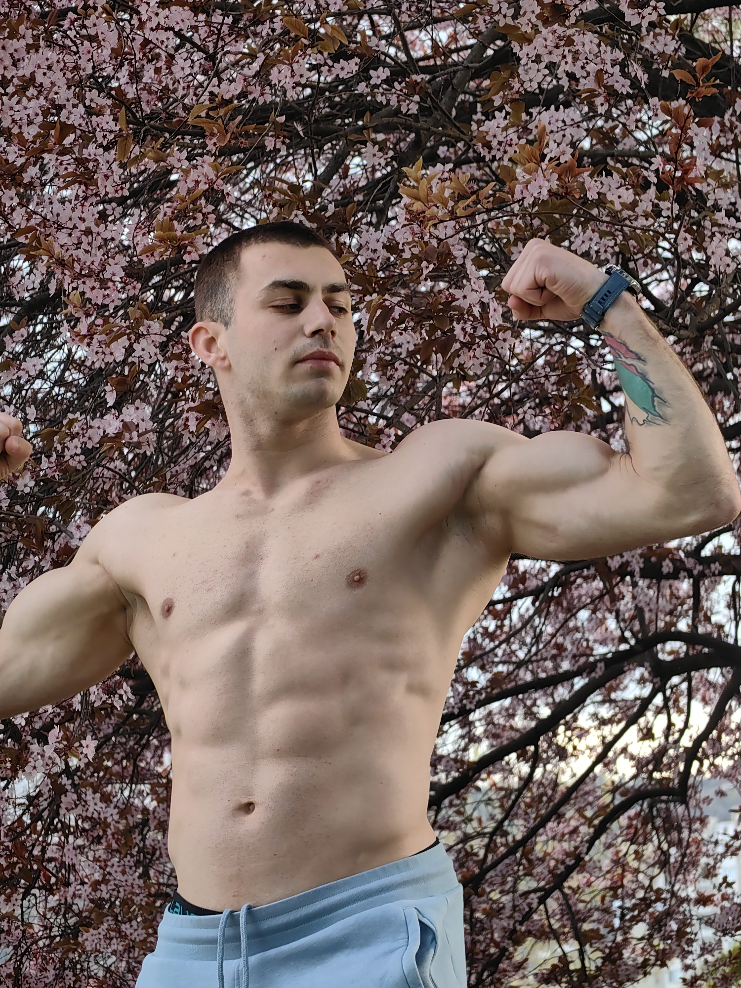 A shirtless, muscular man stands outdoors in front of a tree with pink blossoms. He is flexing both arms, showing off his biceps. The man has short, dark hair, light skin, and is wearing a blue fitness tracker on his left wrist and light blue sweatpants. He has a tattoo on his left arm. The location may be Shumen, Bulgaria.