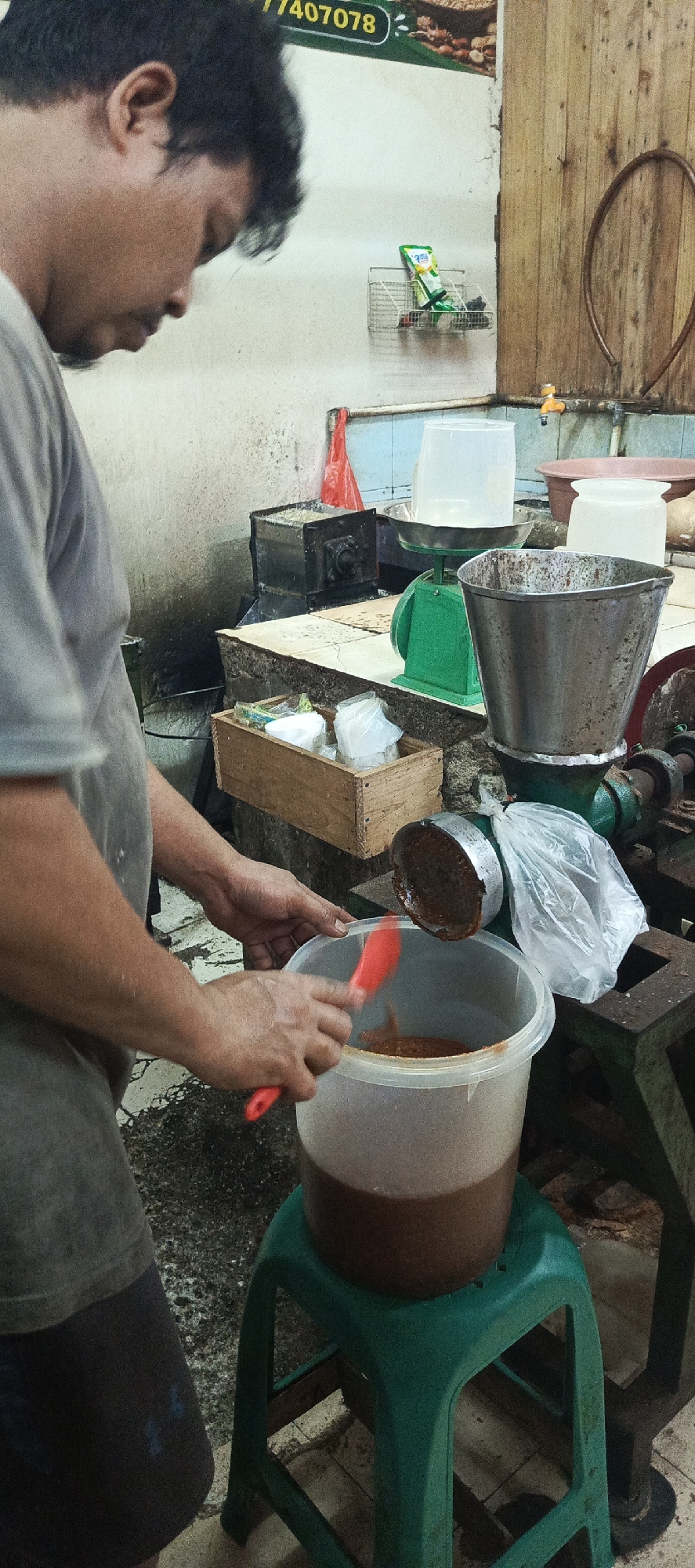 In Sukaresmi, Indonesia, a man with dark hair and fair skin, wearing a grey t-shirt and dark shorts, is standing in front of a green plastic stool. He is holding a red spatula and appears to be transferring a brown, semi-liquid substance from a metal hopper to a large, clear plastic bucket. The bucket contains a significant amount of the same brown substance. Behind him, a green industrial scale is visible with a large clear plastic container on it. To the left of the scale, a black metal machine with visible gears is present. A wooden crate filled with plastic-wrapped items is on a surface to the left of the scale. The environment appears to be an indoor workspace, possibly a food processing area, with concrete floors and a tiled wall. A tap is visible on the tiled wall.