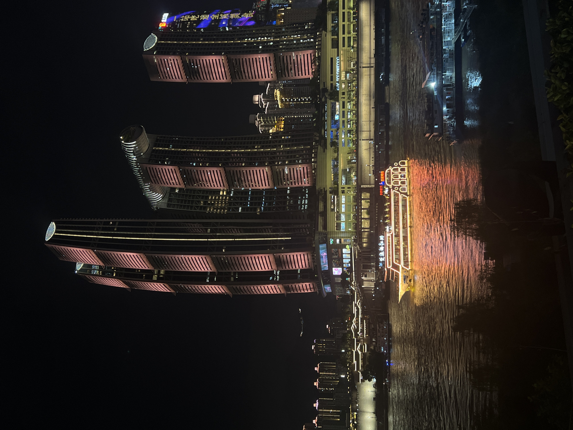 The image depicts a nighttime view of the Jiangbeicheng Subdistrict in China, showcasing a waterfront scene dominated by illuminated skyscrapers and a river with several boats. The foreground shows the dark water of a river with reflections of the lights from the city and the boats. A prominent cruise ship with glowing red and yellow lights is positioned in the center of the frame, on the river. To the right of the cruise ship, another larger boat with numerous lights is docked.
In the background, a cluster of modern skyscrapers rises from the cityscape. The most striking of these are three tall buildings, two of which appear to be connected at a higher level by a structure that curves and is illuminated with bright lights. These buildings are vertically segmented and feature large areas lit in pink and yellow. The skyscraper on the far right has blue and purple lights forming vertical patterns and a curved illuminated top. Further back and to the left, a row of smaller buildings with illuminated windows and roofs are visible, creating a layered urban landscape. The sky is dark and clear, serving as a backdrop for the illuminated city.
