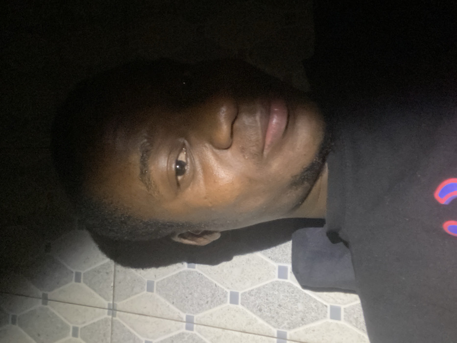 A close-up shot of a dark-skinned man laying down. The man is on a tiled floor with a repeating pattern. He is wearing a black shirt with a blurred design on the right shoulder. His head is slightly tilted, facing the camera with his eyes partially open. The lighting is low, casting shadows on his face and hair, which is short and closely cropped.
