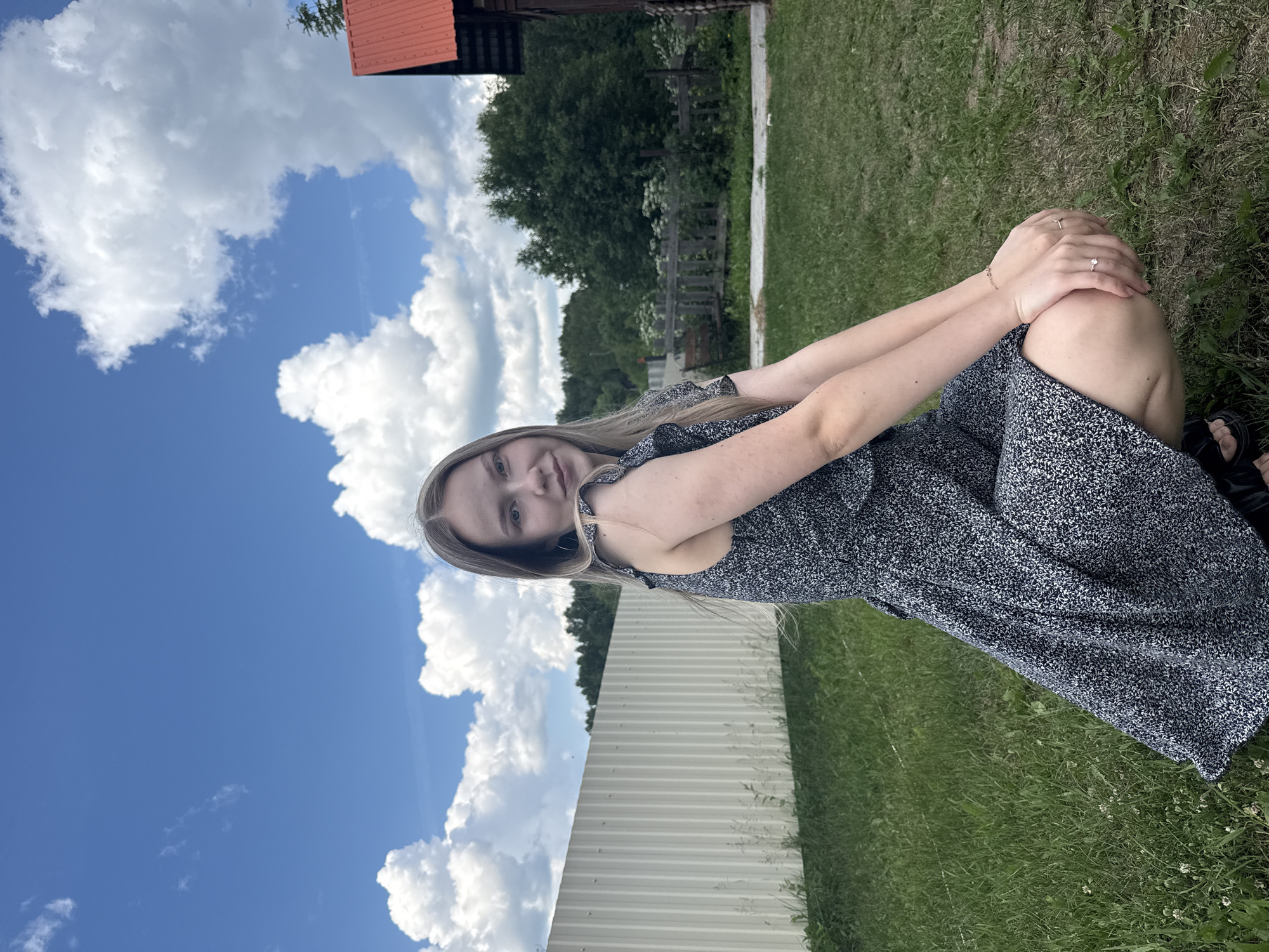 A young woman with long blonde hair is crouching in a grassy area. She is wearing a dark, floral-patterned dress and black sandals. Her hands are resting on her knees. Behind her, a white corrugated fence runs horizontally across the frame. In the distance, trees and a wooden structure with a red roof are visible. The sky is blue with scattered white clouds. The scene appears to be outdoors, likely in a rural or suburban setting, possibly in Zhigalki, Russia, given the context provided.