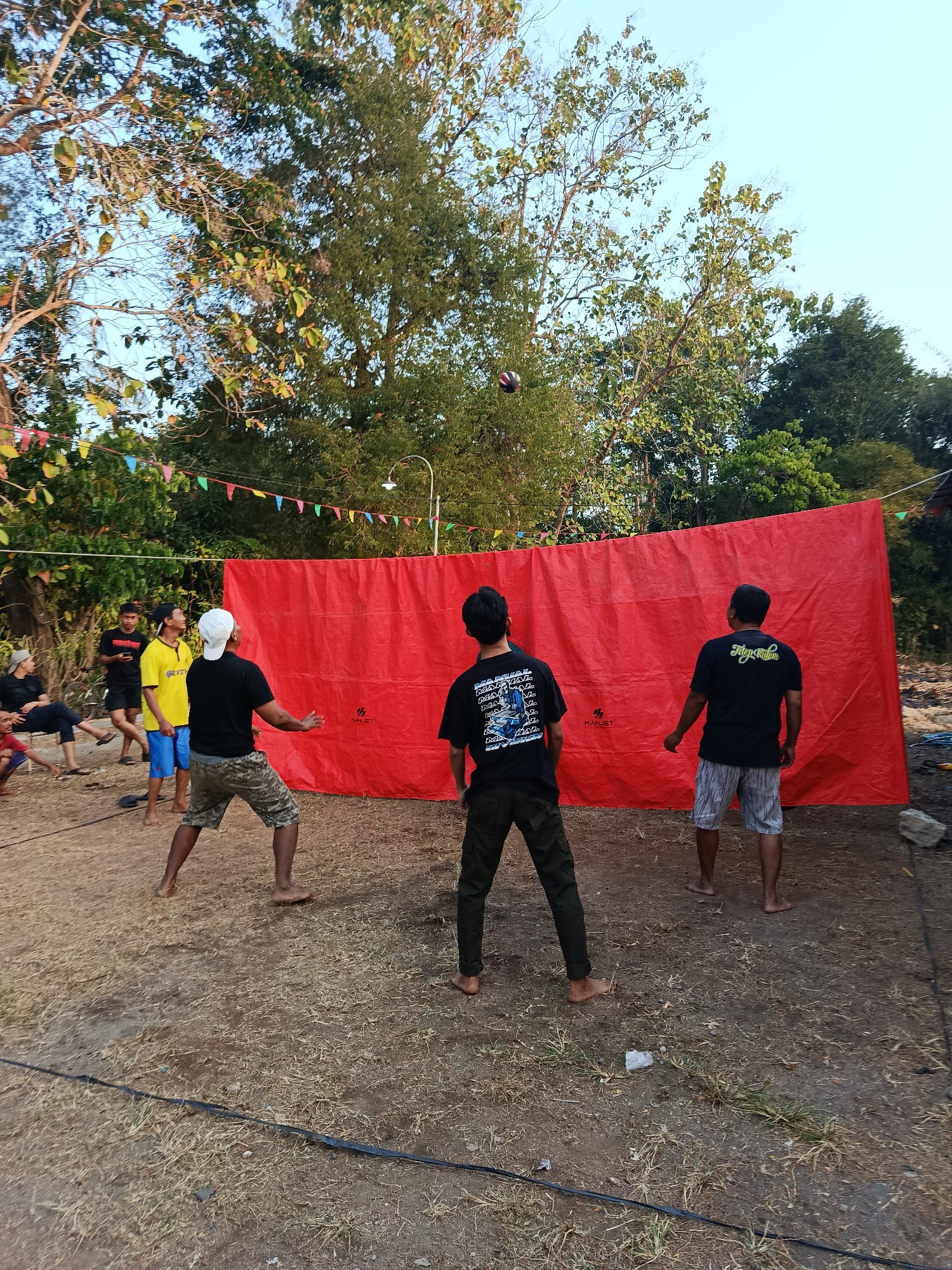 A group of young men are playing volleyball in an outdoor setting in Sedayu, Indonesia. The game appears to be casual, with players dressed in t-shirts and shorts, some barefoot. A large red tarp is stretched out, likely serving as a makeshift net or backdrop for the game. Trees and foliage surround the area, suggesting a rural or park-like environment. Decorative flags are strung up in the background, adding a festive touch to the scene. A volleyball is visible in mid-air, indicating the game is in progress.