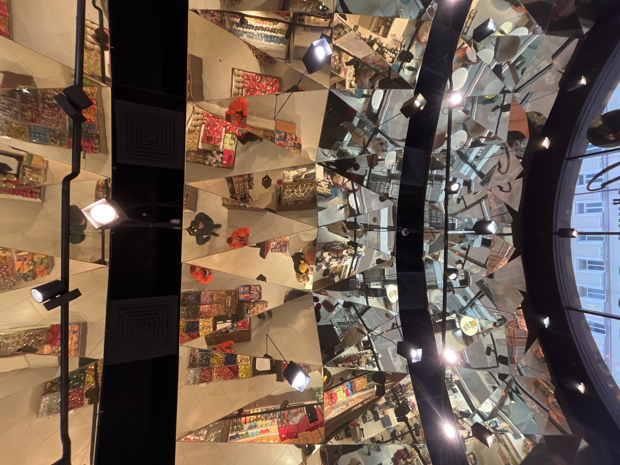 The photograph depicts a complex mirrored ceiling installation in a retail environment, likely a department store in Moscow, Russia. The ceiling consists of many small, irregularly shaped mirrors, creating kaleidoscopic reflections of the store's interior. Visible within the mirrored fragments are shelves stocked with various goods, notably candies and sweets, as well as reflections of shoppers. The ceiling also includes structural elements such as black beams and lighting fixtures, some of which are directed towards the mirrored surfaces, enhancing the reflections. Through a curved window in the upper right, the exterior architecture of a building can be seen.