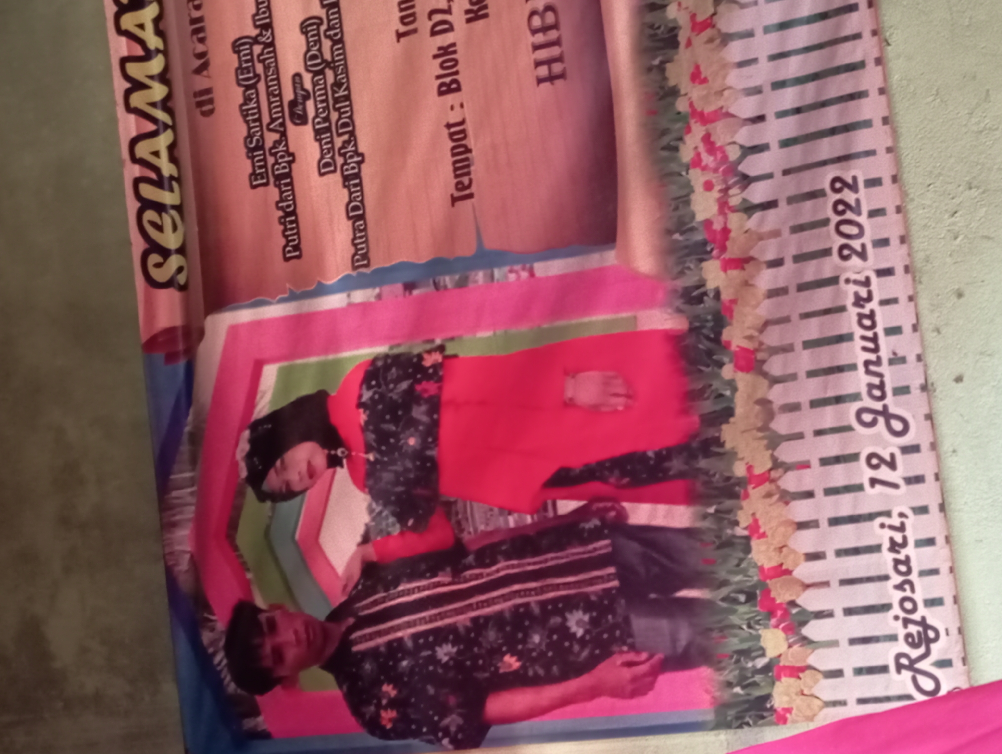 The image is a photograph of a banner with text and images. At the bottom of the banner, text reads "Rejosari, 12 Januari 2022", indicating the location and date. The banner features a picture of a man and a woman, presumably a couple, dressed in traditional attire. The man is wearing a patterned shirt and jeans, while the woman is wearing a hijab and a red outfit over a patterned dress. The background of the banner includes decorative elements like a white picket fence and flowers, along with text that appears to be an announcement for an event, possibly a wedding. The overall tone suggests a celebration or formal occasion.
