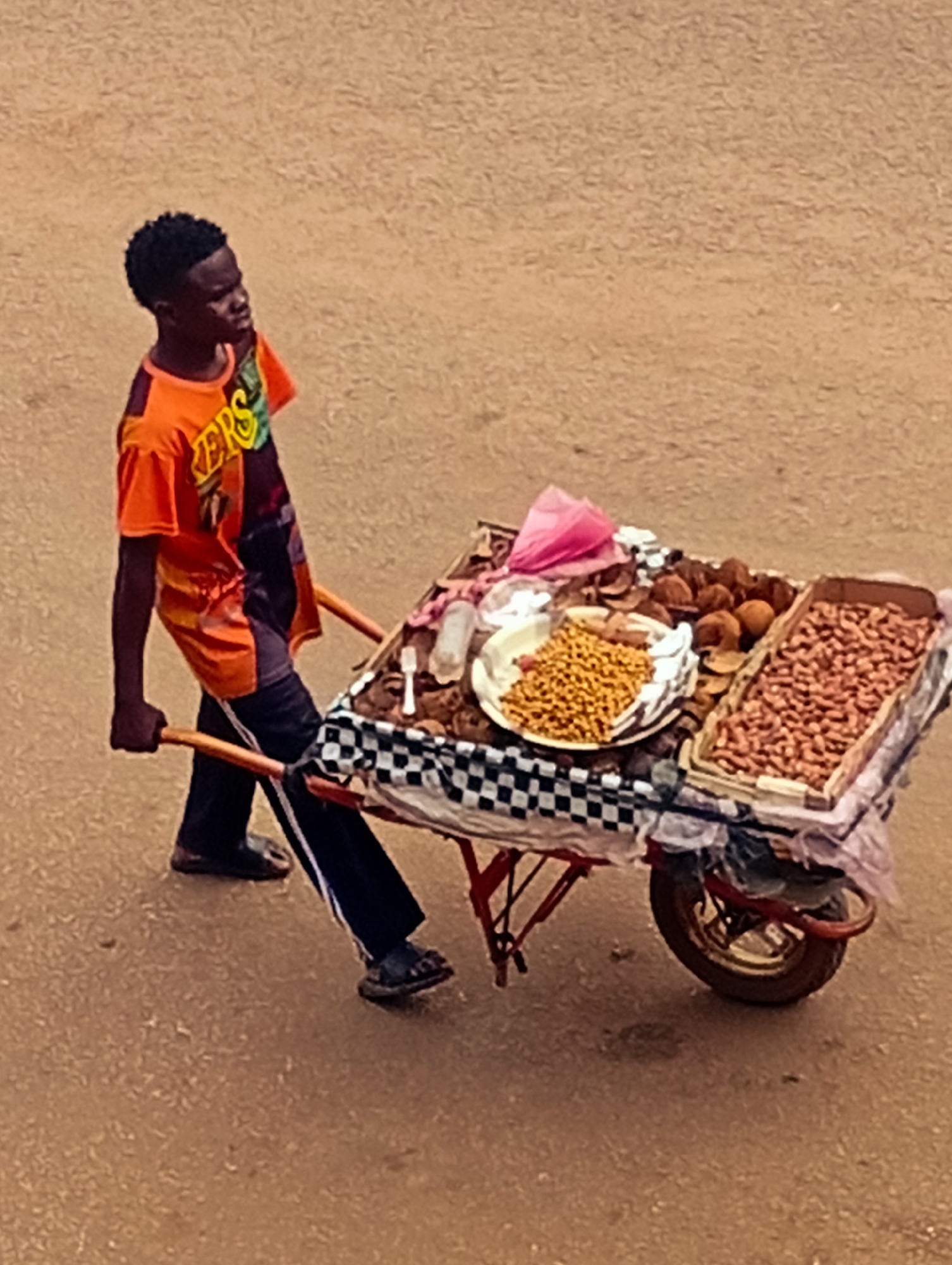 A young man in Bukuru, Nigeria, pushes a red-framed, single-wheeled cart laden with food items for sale. The cart, covered with a checkered cloth, holds a tray of peanuts, a plate of yellow snacks, and several round brown fruits or nuts. The man wears an orange and black t-shirt and black pants with a white stripe. He is walking on a tan-colored surface.