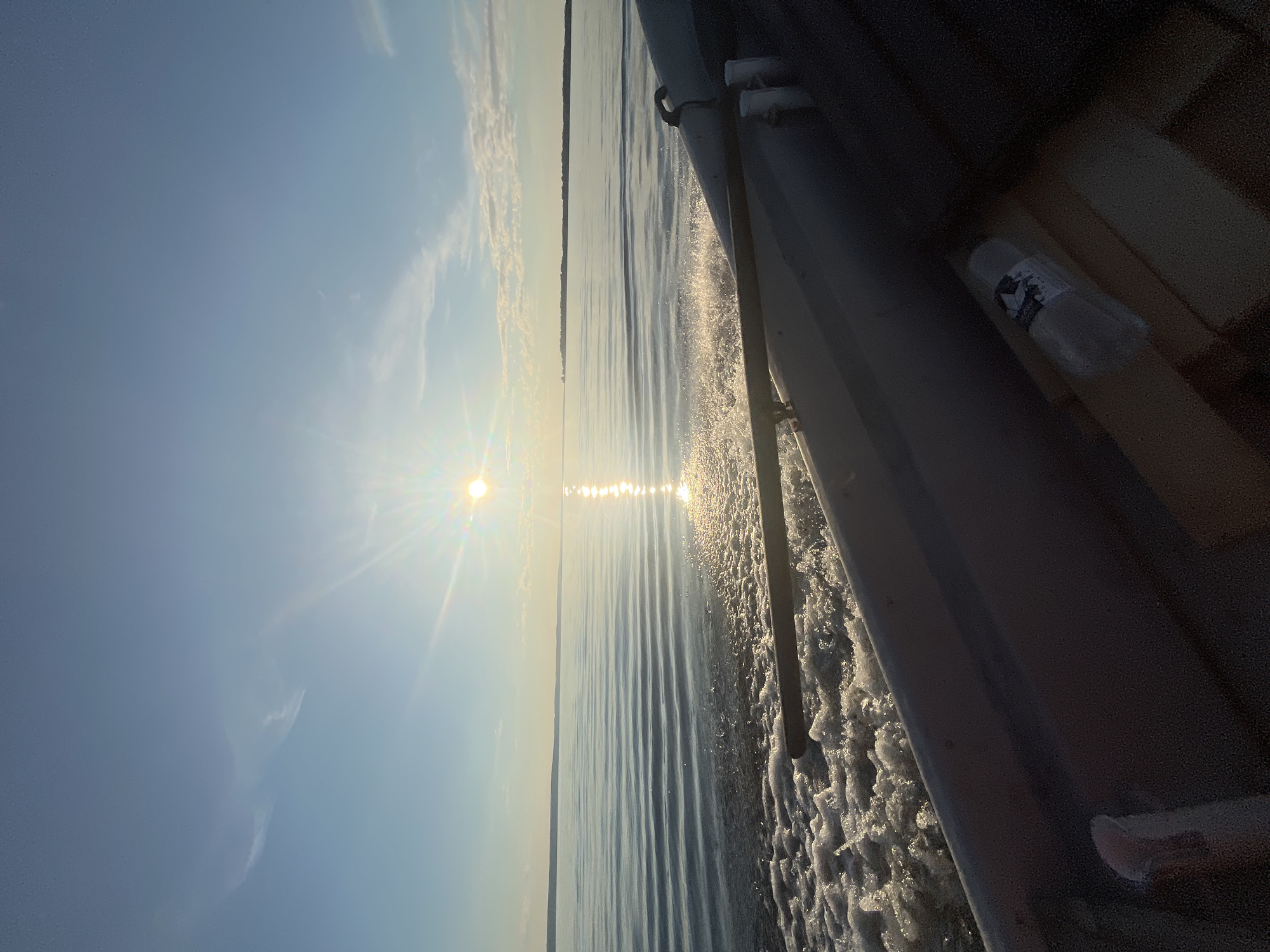 The image shows a serene water scene, likely taken from a boat moving through calm waters. The sun is low in the sky, casting a bright reflection on the water's surface and creating a shimmering path. Wispy clouds are scattered across a clear blue sky. In the foreground, a portion of a boat's interior is visible, including a grey railing and some wooden surfaces. A plastic water bottle with a label is also present inside the boat. The distant horizon shows a faint outline of land. The scene suggests a peaceful moment of travel on a body of water, possibly a lake or a calm sea, under a bright sky. The location is identified as Siukterka, Russia.